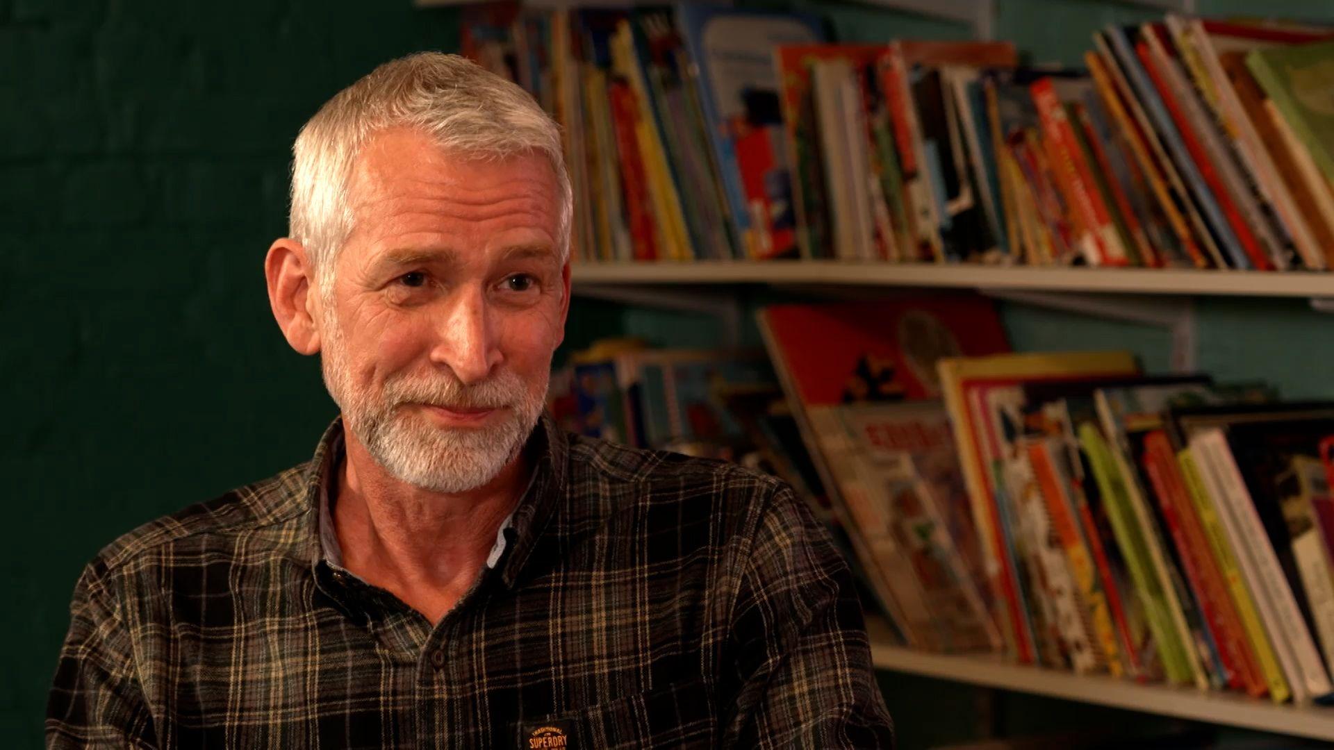 A man with white hair and a beard, Adam Hargreaves, sits in front of a shelf of books