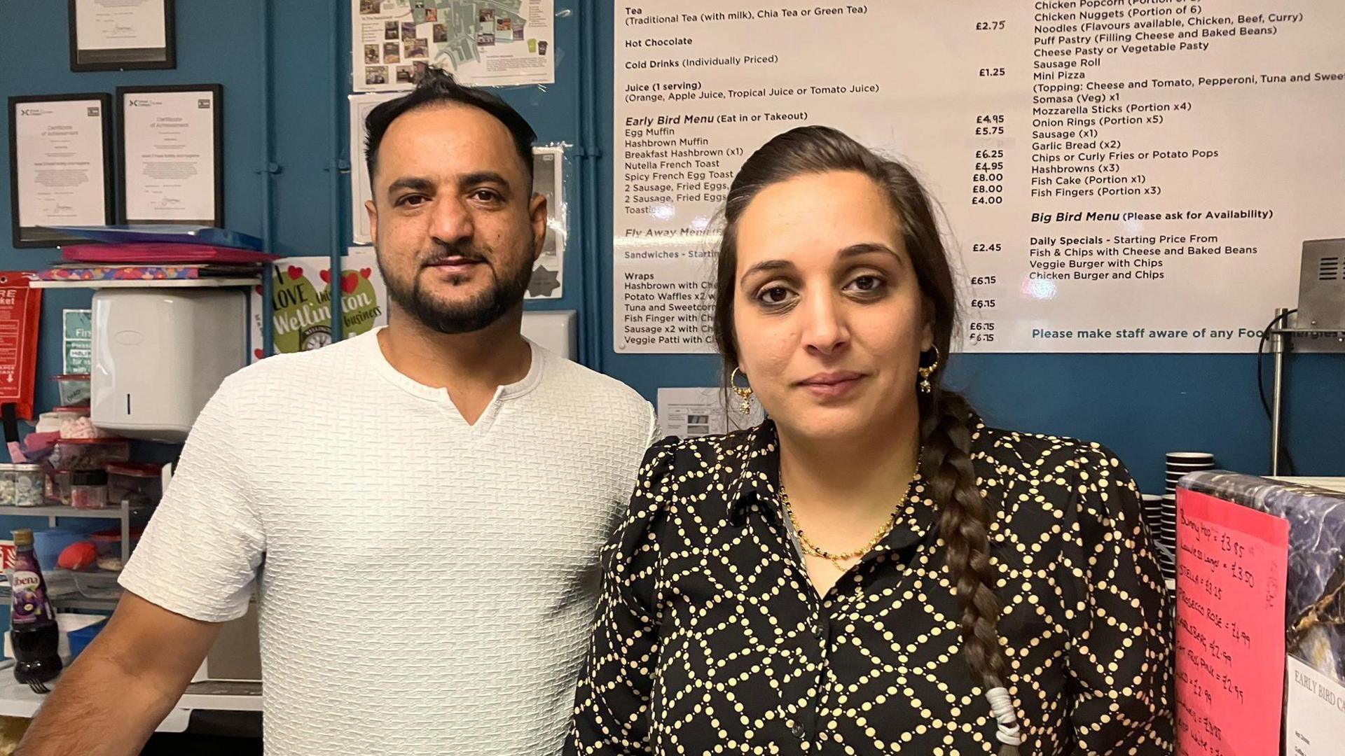 A man in a white t-shirt stand next to a woman with dark plaited hair, wearing a black and yellow patterned shirt. They stand behind the counter of a cafe, which has a large menu up on a board on the wall behind them.