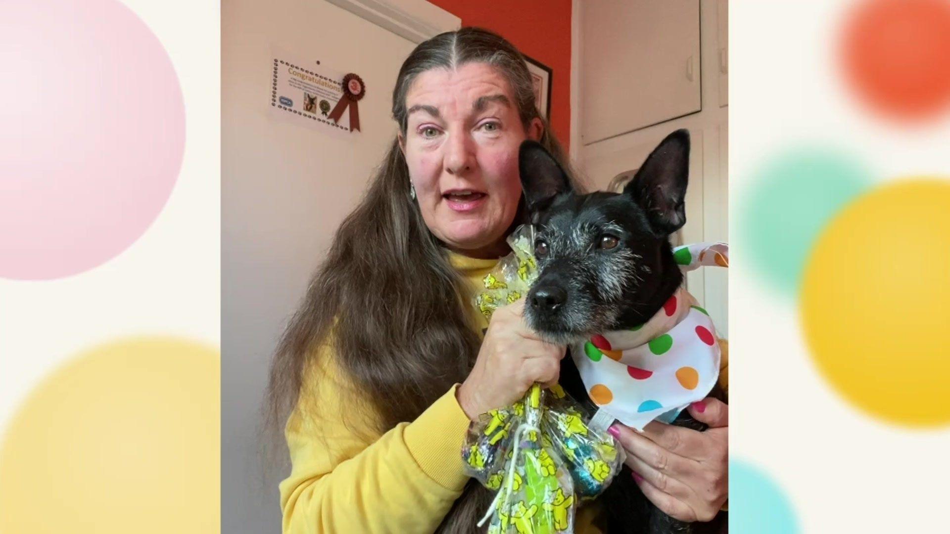 A woman holding a dog wearing a spotty and white bandanna around its neck. The woman is also holding sweets in packets.