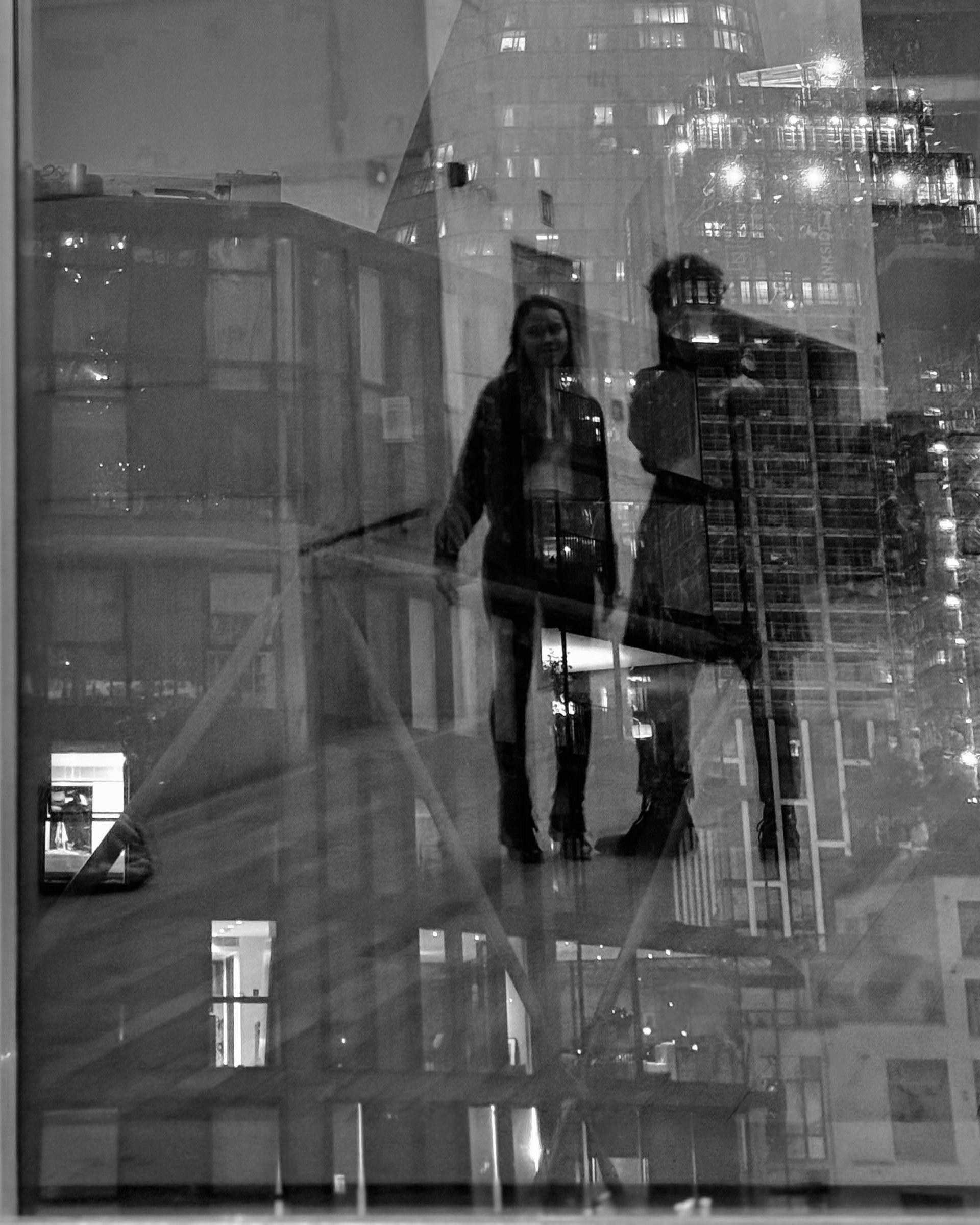 A black and white image of two figures - a man and a woman - reflected in glass windows. They are in a cityscape.