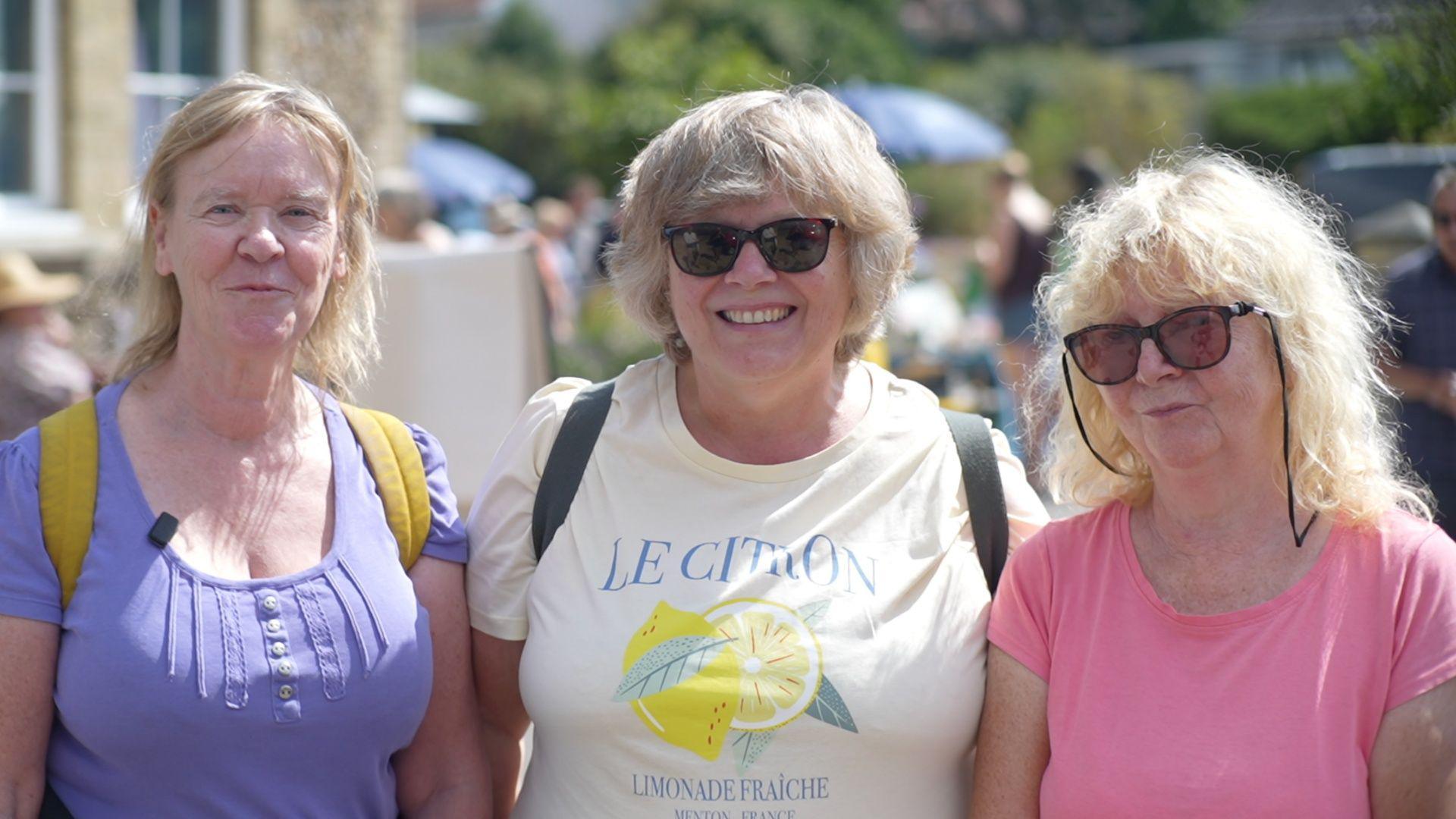 Lynne Sawyer,  Debbie Saxby and Carol Alder, who are standing outside Westleton Village Hall. They are all smiling. Lynne is wearing a purple T-shirt, Debbie a white T-shirt and Carol a pink T-shirt.