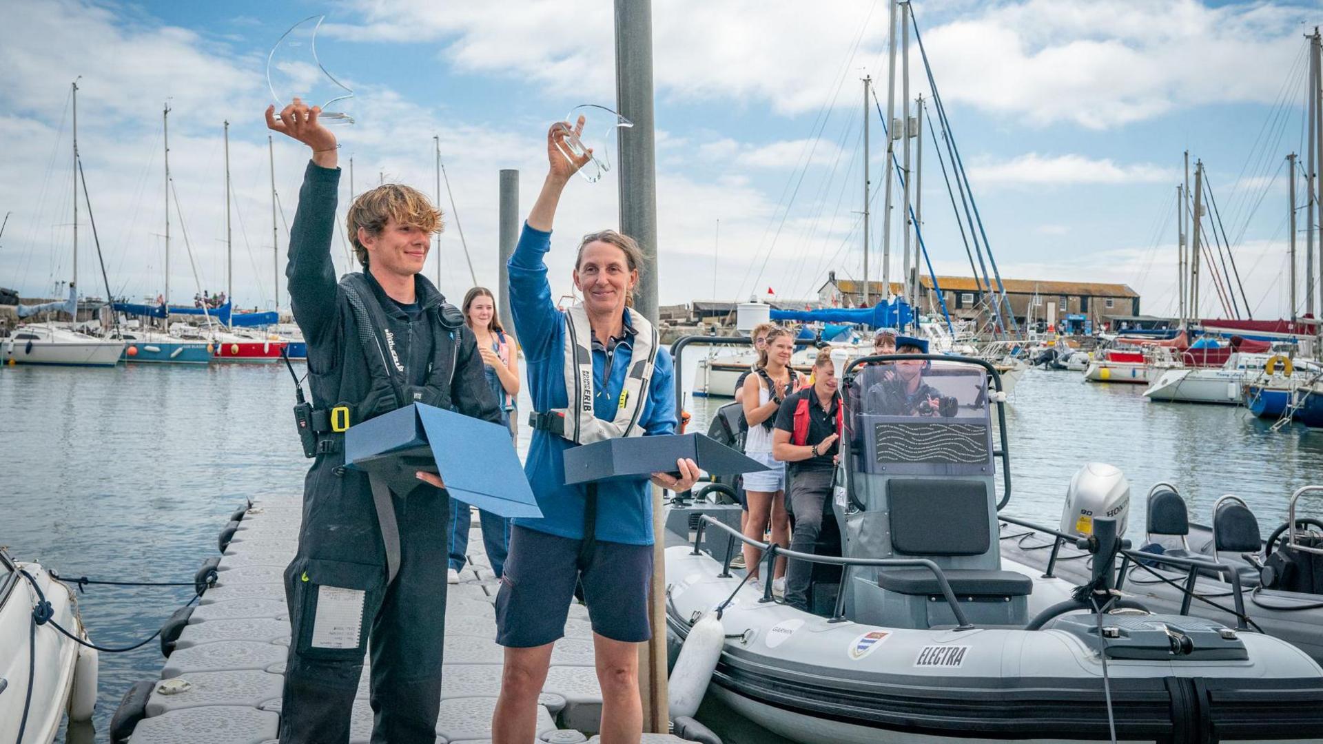 Harry Besley and Project Manager Jaqui Besley wearing life jackets and holding glass trophies shaped like waves in the air. Behind them is the Lyme Regis harbour, full of electric boats and sailboats