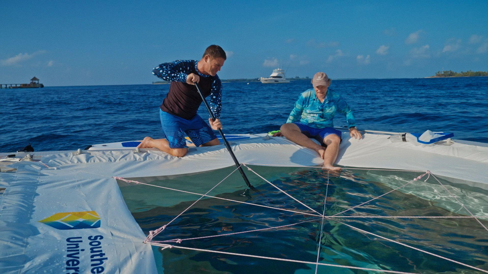 Steve Backshall and Prof Peter Harrison sitting on the edge of an inflatable pool in the ocean. Steve is holding a net on a long pole, dipping it into the water.