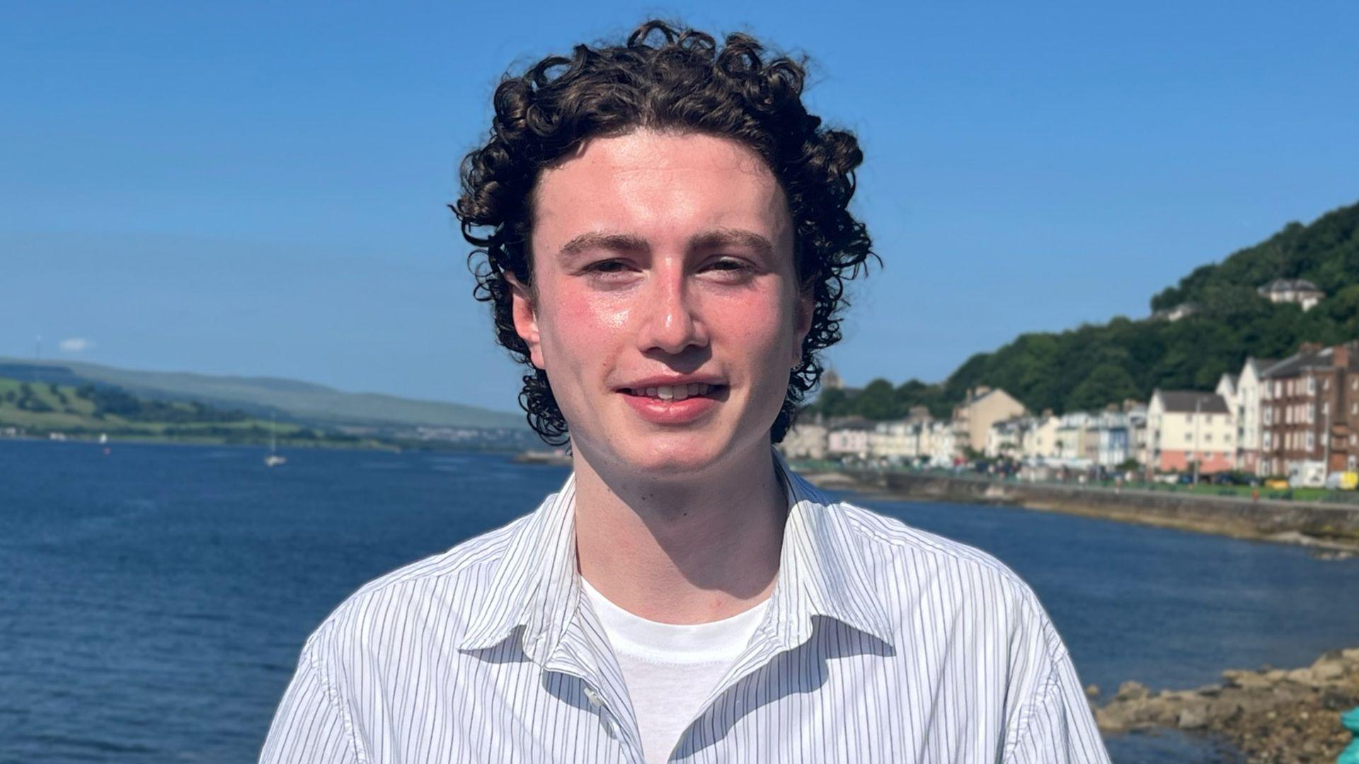 Matthew Quinn stands by a waterfront with calm bluish water, coastal buildings and greenish hills down him.