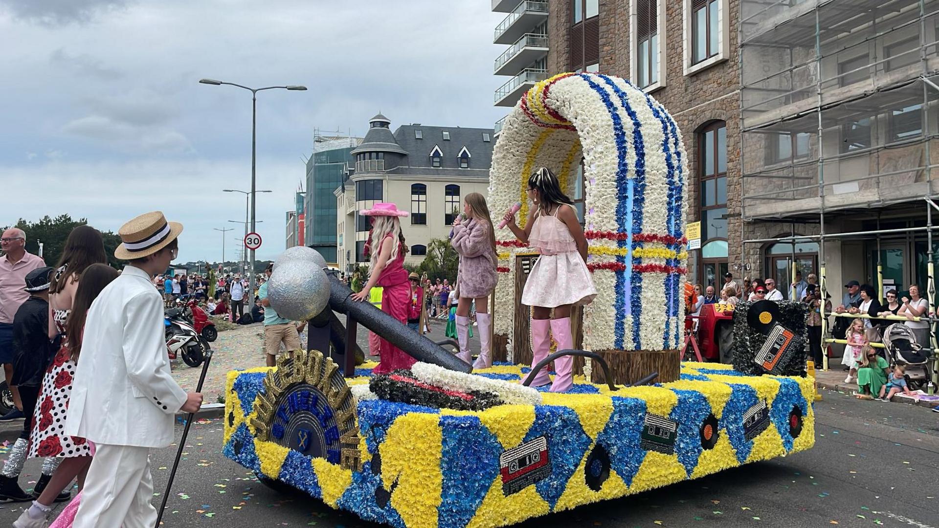 A float designed to look like a jukebox. There are three girls holding microphones. One is wearing a pink cowgirl outfit and the other two are wearing short dresses and long pink boots. The float features two giant silver microphones and the lettering: JUKEBOX JIVE. Two boys wearing suits and hats walk behind the float along with a girl in a 60s style dress. People are lined up on the streets watching.