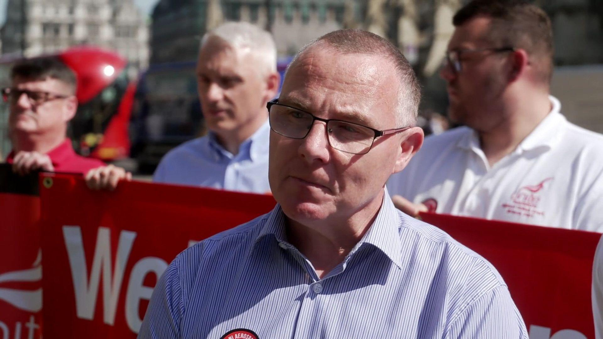 A bald man in a striped blue and white shirt, wearing glasses, stood in front of protesters holding a red banner