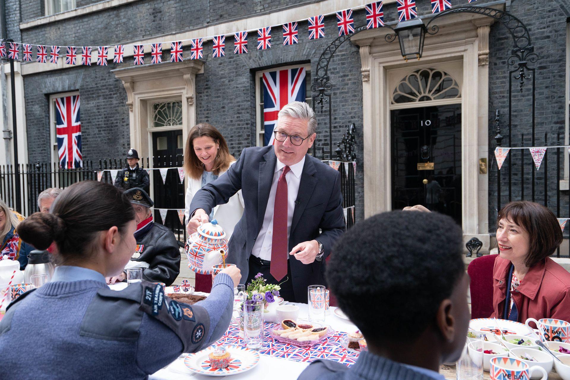 Keir Starmer apprehensively pours a teapot into a teacup held by a person wearing military uniform, with other attendees around the table that contains various bits of afternoon tea in front of Number 10 Downing Street. Union flag bunting adorns the building.
