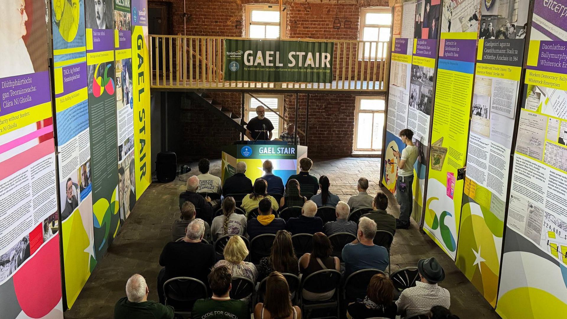 A man stands at a podium in front of an audience of about 30 people. He is in a hall where, to the left and right, there are long, very tall displays for an exhibition. 