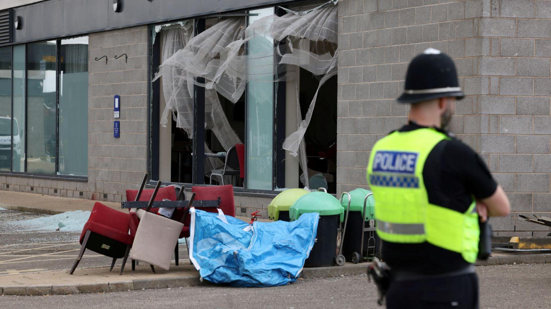 A police officer in a hi-vis vest stands with his back turned and arms folded in front of a building. Furniture and broken glass can be seen piled in front of several broken windows.