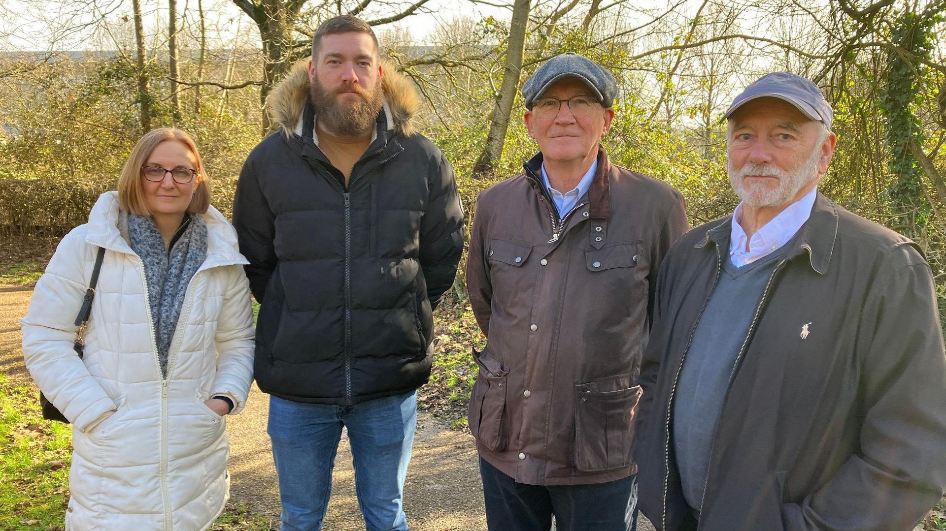 A picture of four people stood on a path in front of where a recycling centre could be built in Milton Keynes. There is a woman with shortish brown hair wearing a white coat, a man with short hair and a full beard with a black coat, then two men with caps on, one is clean shaven but wearing a brown coat, the other has a white beard and has his hands in his pocket.