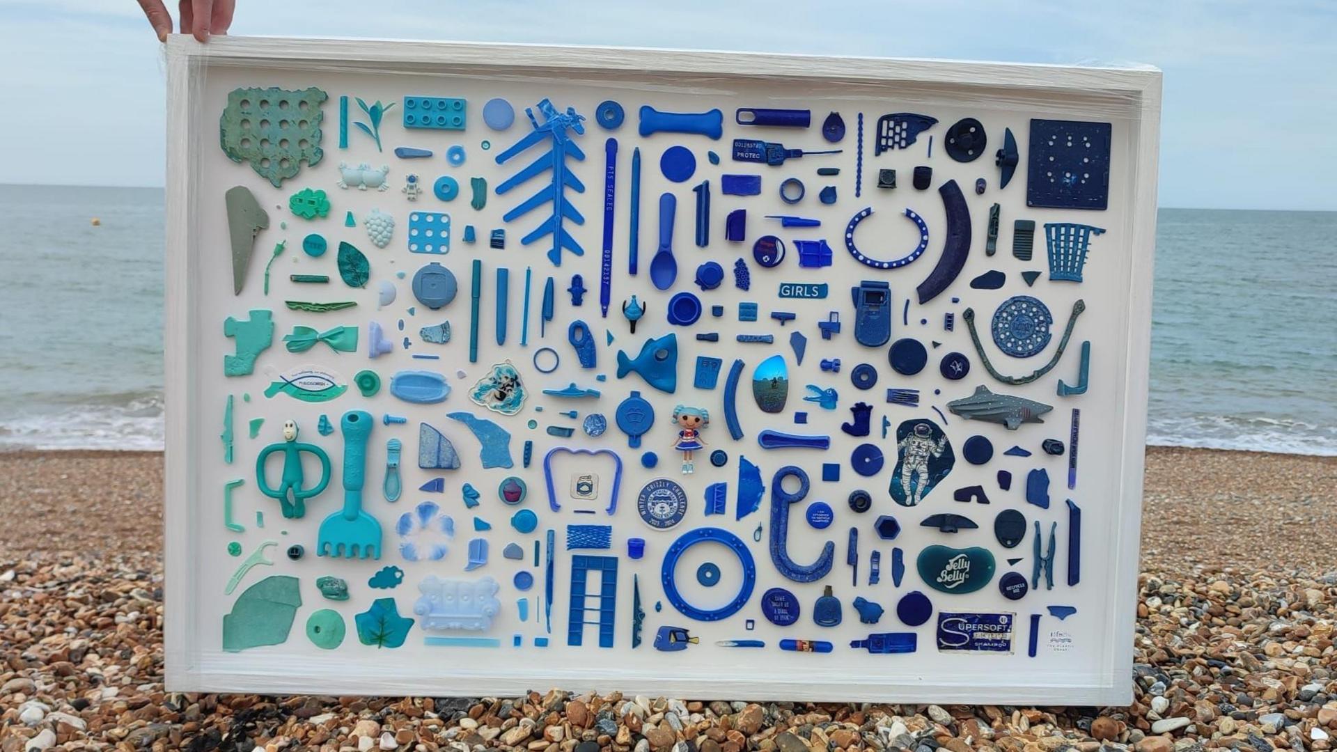 Various shade of blue sea plastic arranged and stuck to a white framed background. A hand is holding the top left of the frame to keep it upright. It is on the pebbles of a beach. The sea is in the background