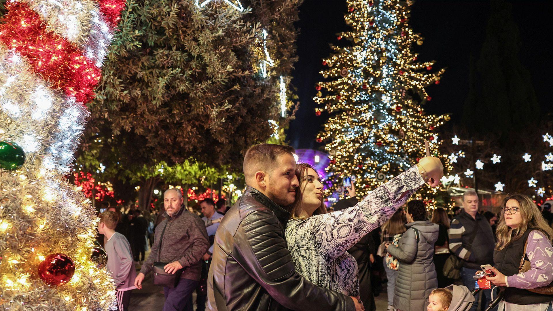 a man and a woman pose as the woman takes a selfie and crowds walk past - in the foreground is red and white striped candy cane style tinsel and in the background is a christmas tree covered in warm and cool white lights.