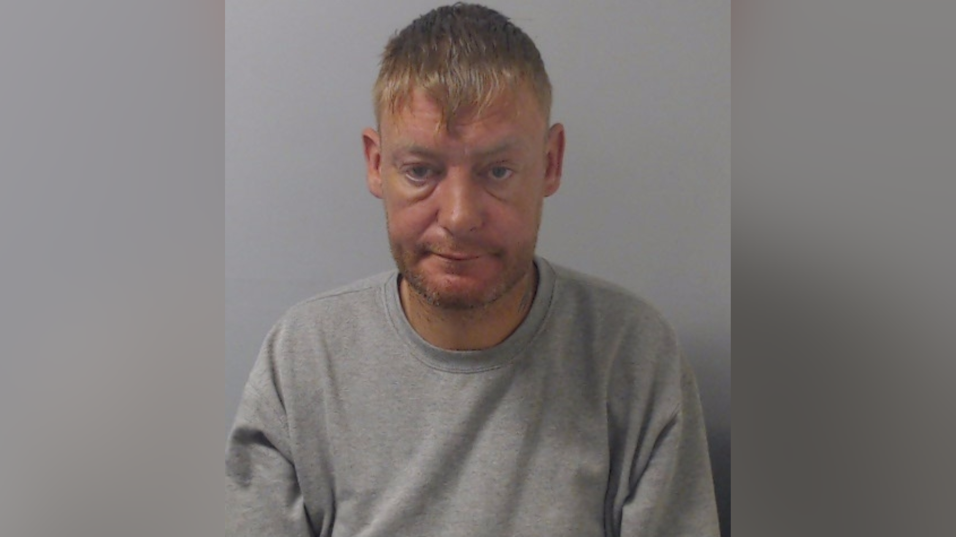 An image of a man, wearing a grey jumper. He is looking at the camera. He has short blonde hair.