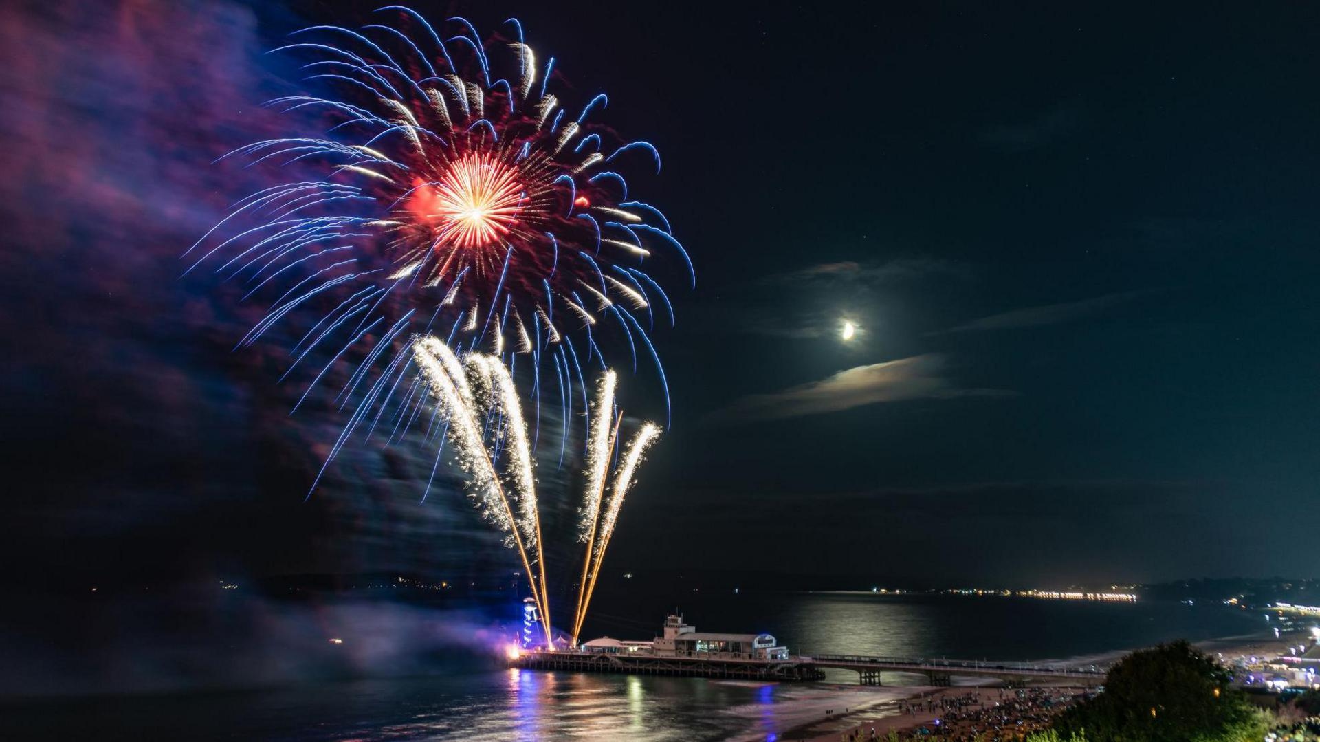 Large fireworks are exploding over Bournemouth pier at night. The moon is visible with hundreds of people on the beach.