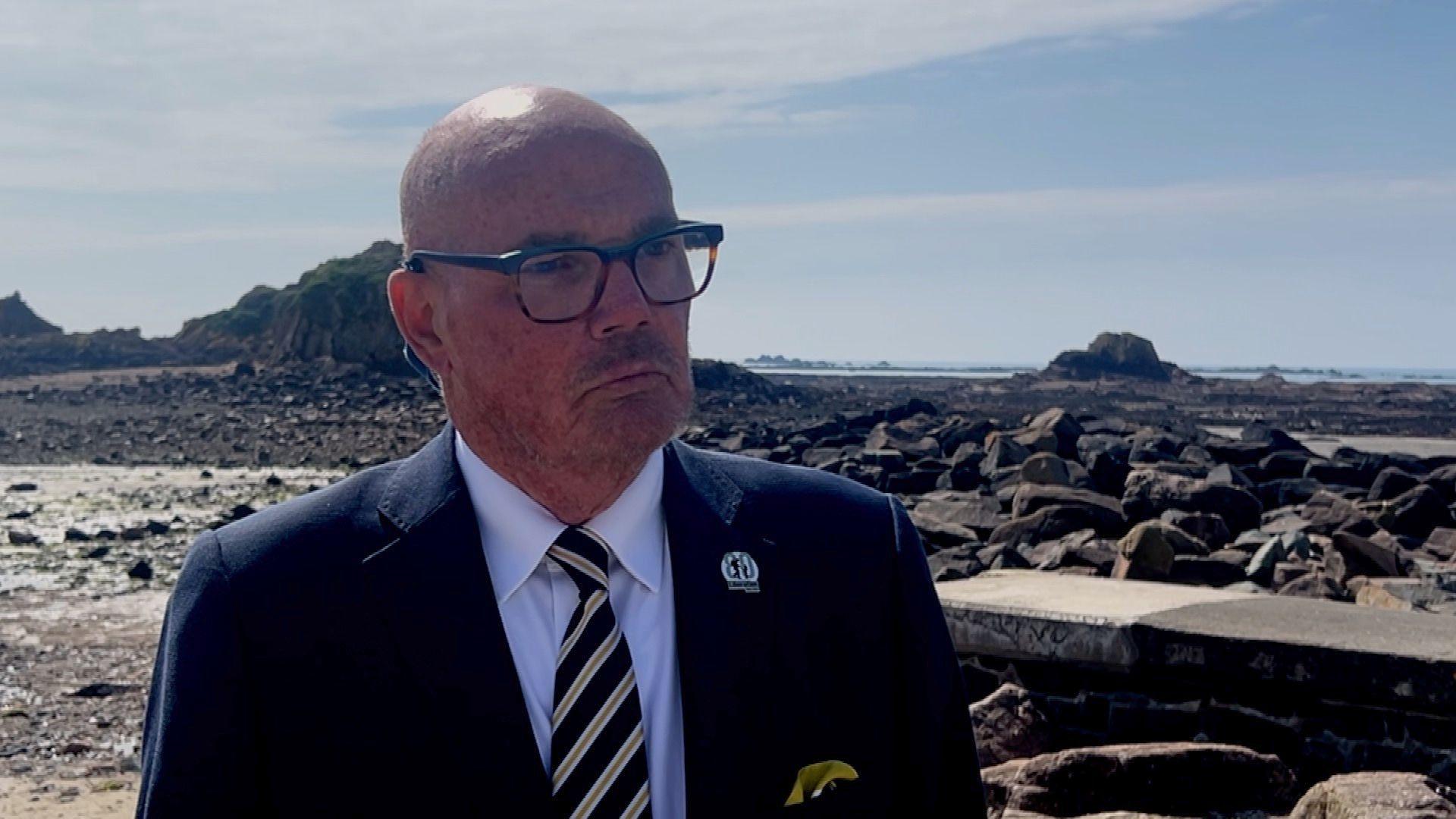 Constable Marcus Troy is wearing a suit and glasses. He is stood on St Clements bay. Behind him is the sea and lots of rocks.
