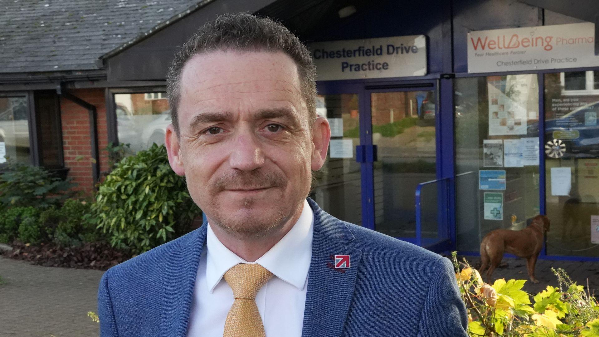 Ian Fisher smiles at the camera outside a GP medical practice. He has short dark hair and wears a blue suit, white shirt and a gold tie. Some bushes can be seen around him outside the GP.