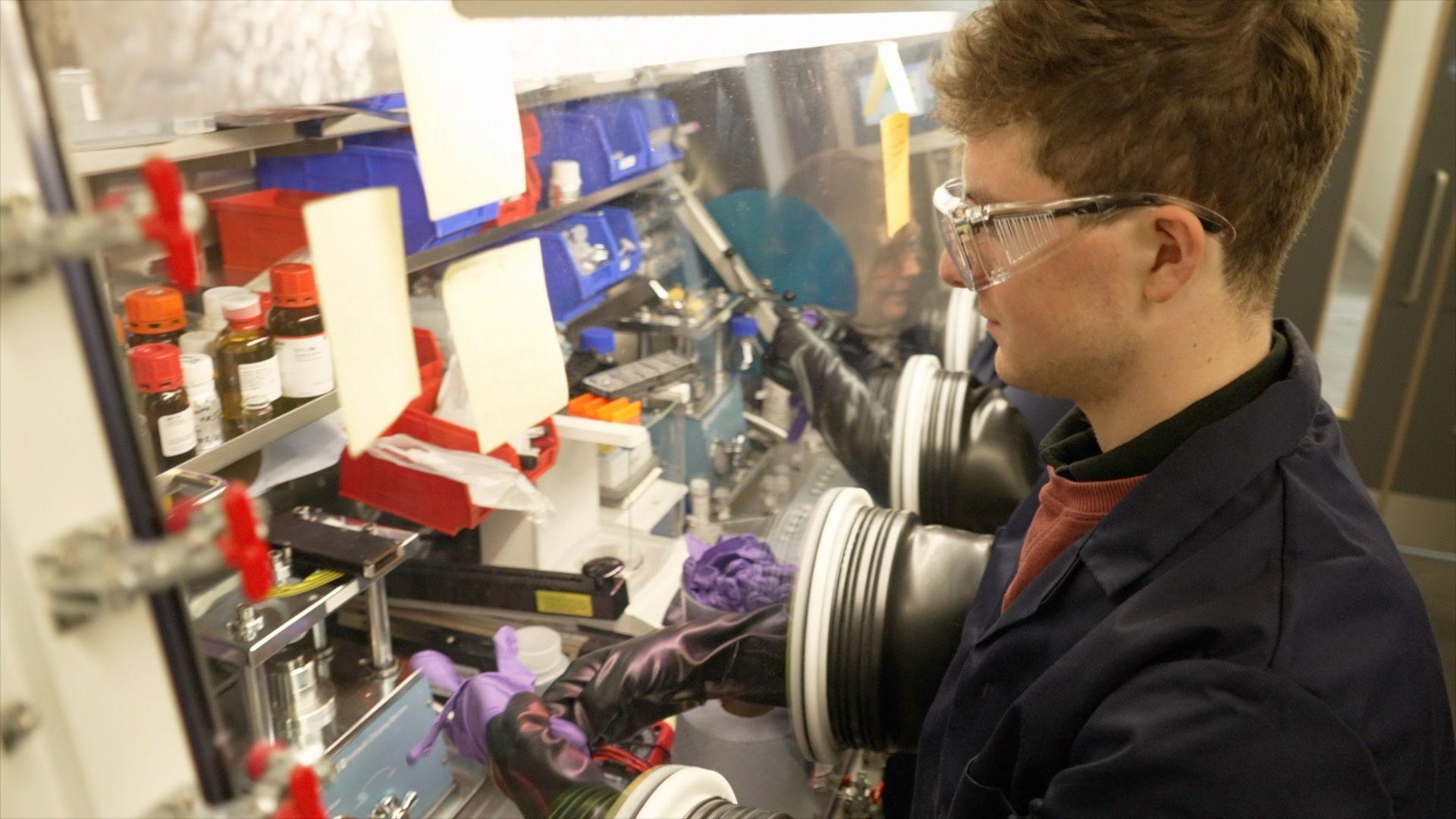 A researcher in a black lab coat is conducting experiments. His arms are inside long rubber gloves which pass through two round portals into a sealed glass lol chamber. It allows him to work with materials inside the chamber without being exposed to them. The chamber contains shelves covered in red-capped bottles, jars and equipment. Hand-written notes are stuck to the exterior of the glass. 