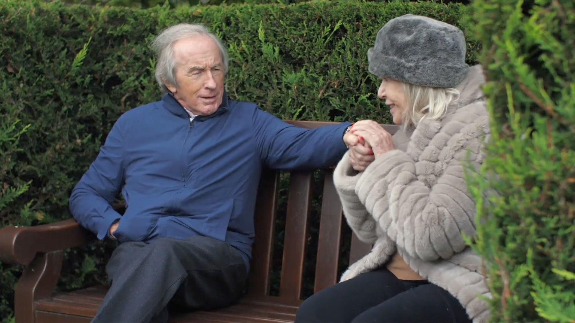 Sir Jackie Stewart sits next to his wife Lady Helen Stewart on a bench at their home. She is wearing a tawny fur type coat and grey fur style hat. She is caressing her husband's hand and smiling at him, he is holding out his hand and smiling at her. His other hand is in his pocket.