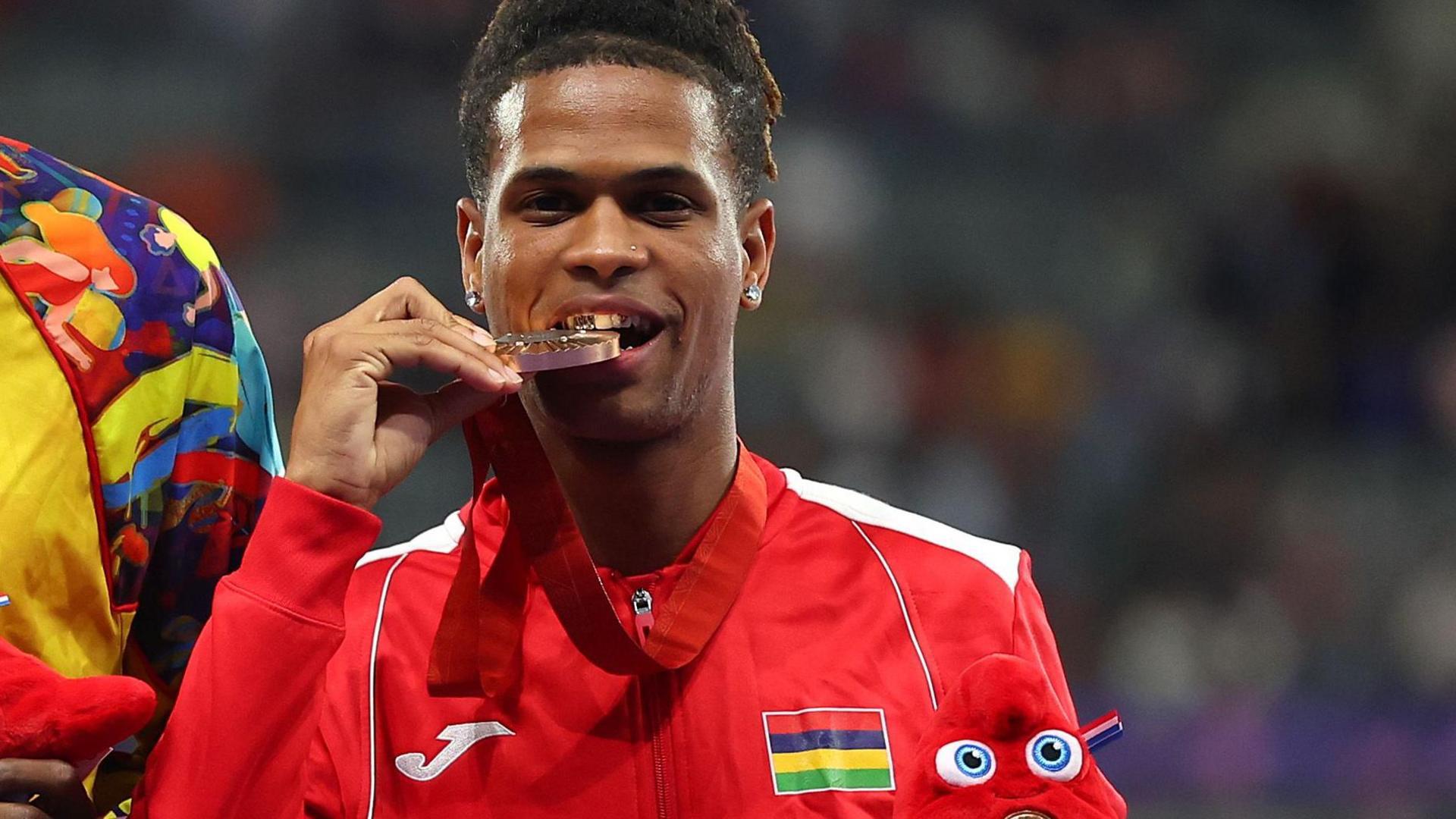 Yovanni Philippe bites down on a bronze medal as he celebrates on the podium at Paris 2024 while wearing a red Mauritius tracksuit