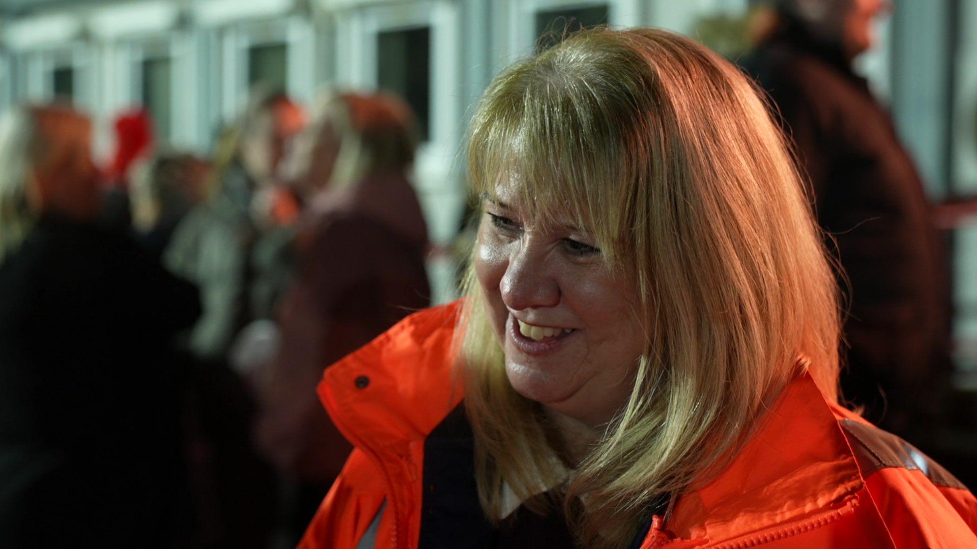 A woman with long blonde hair and fringe smiles wearing an orange high-visibility jacket.