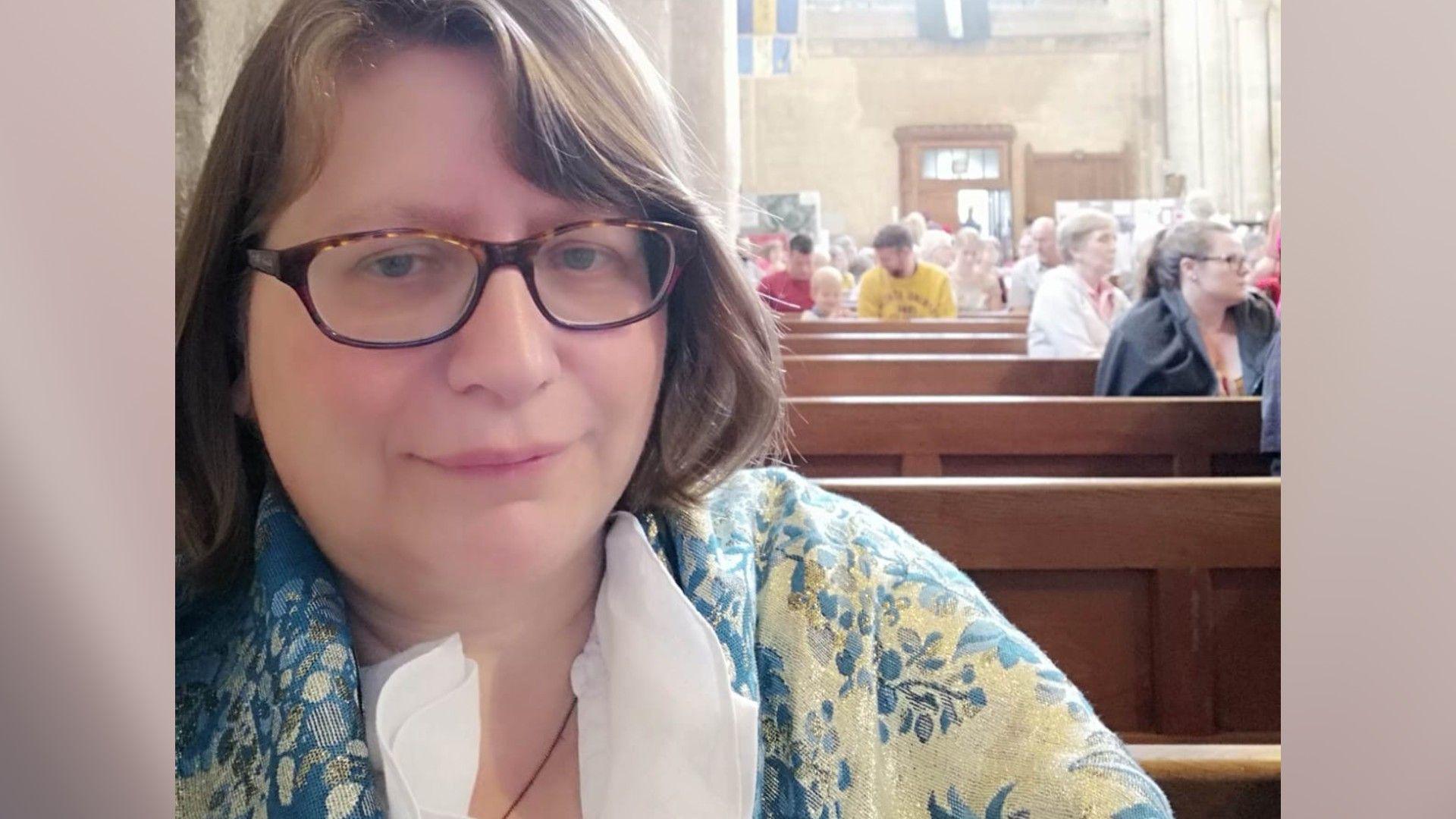 A woman sitting inside Thorney Abbey wearing a floral cardigan, smiling for a selfie.