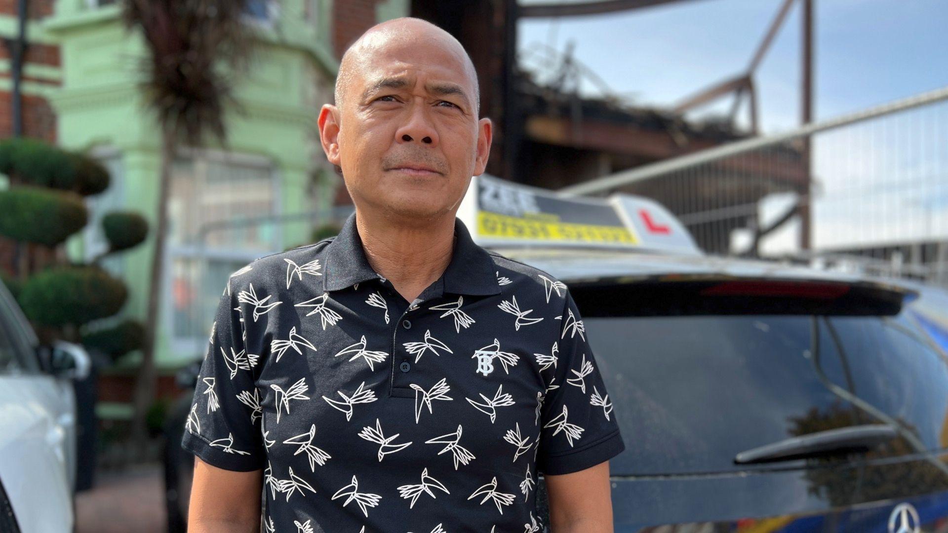 A man looking straight at the camera while wearing a black polo shirt with a white pattern on it. He is standing outside his home, in front of a black car.
