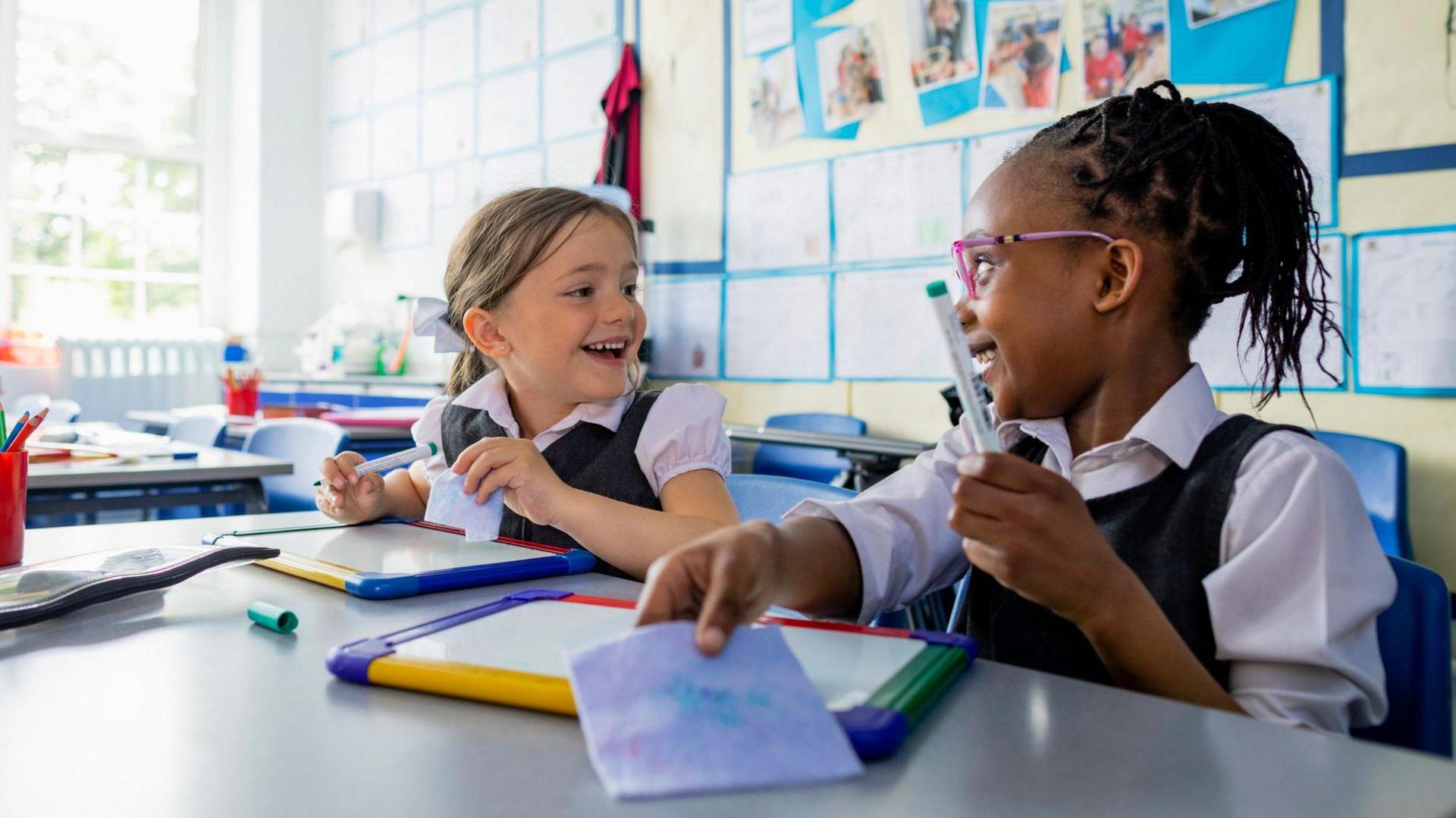 Two children in a classroom with whiteboards and pens, smiling at each other