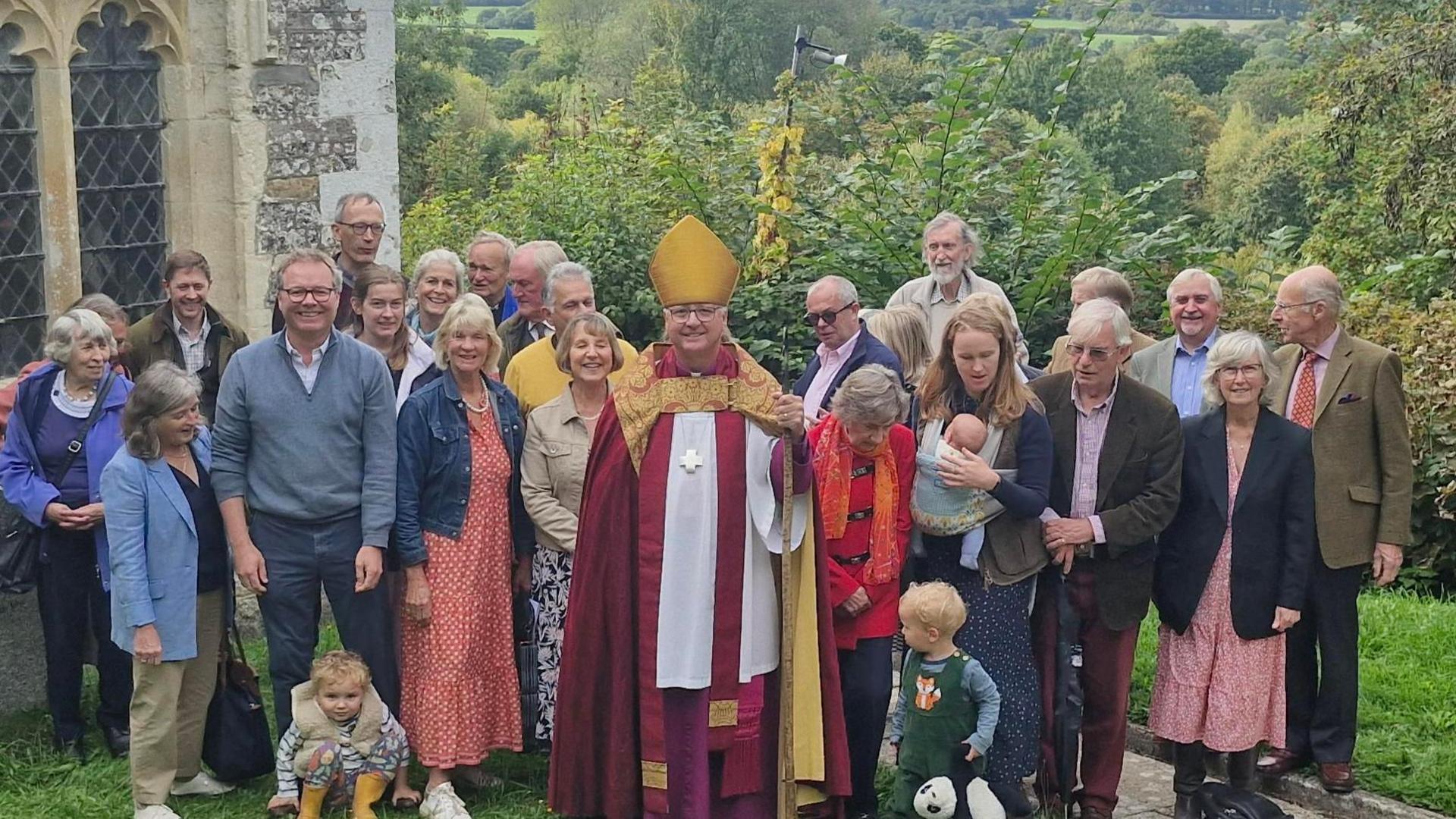 A 'team' photo of parishioners and the Bishop of Salisbury in gold and maroon robes, in front of a corner of the church, overlooking countryside below