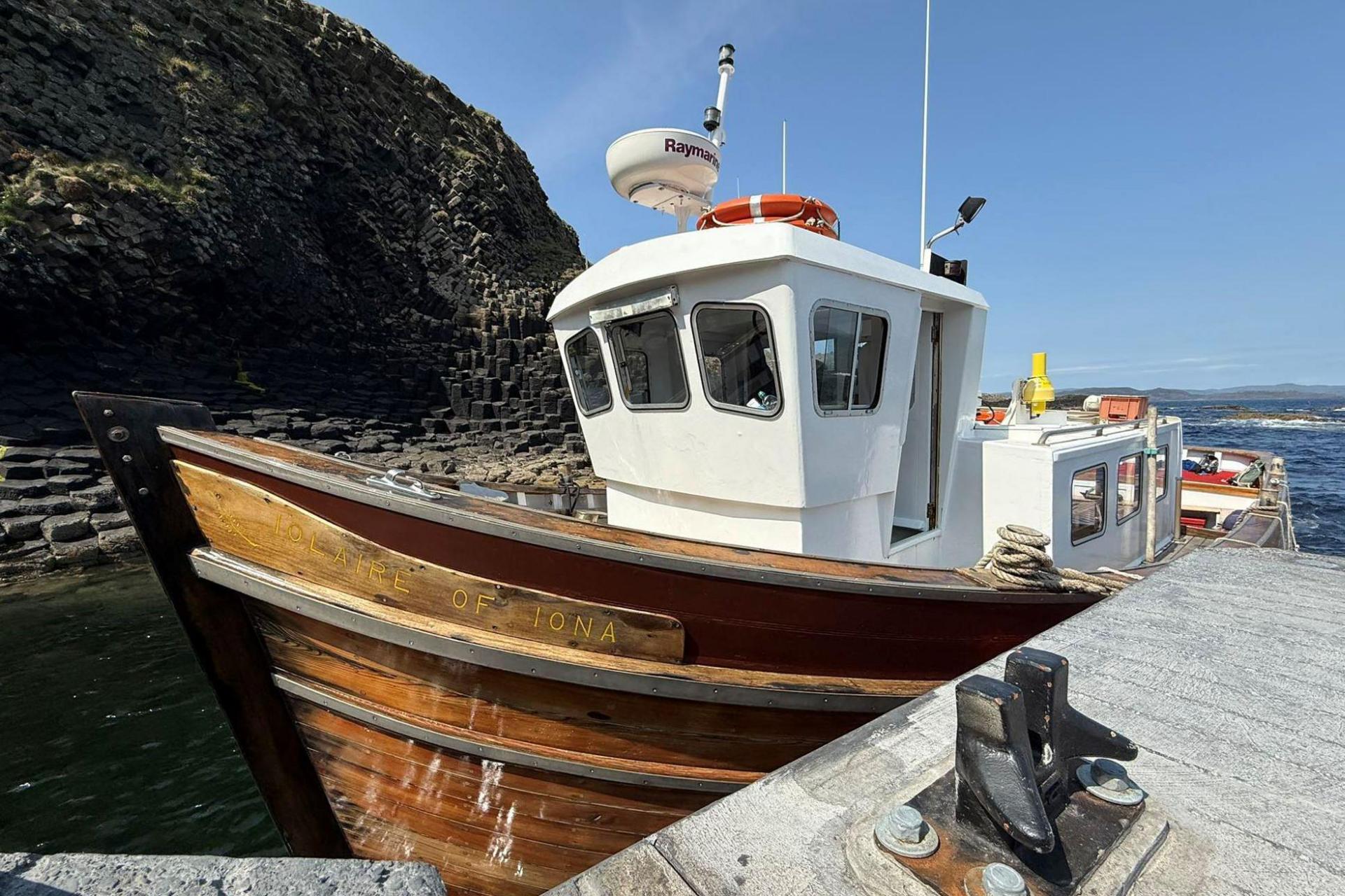 A small boat with a wooden hull and a white cabin tied up at the new jetty. In the background, a cliff of hexagonal rock rises from the sea.