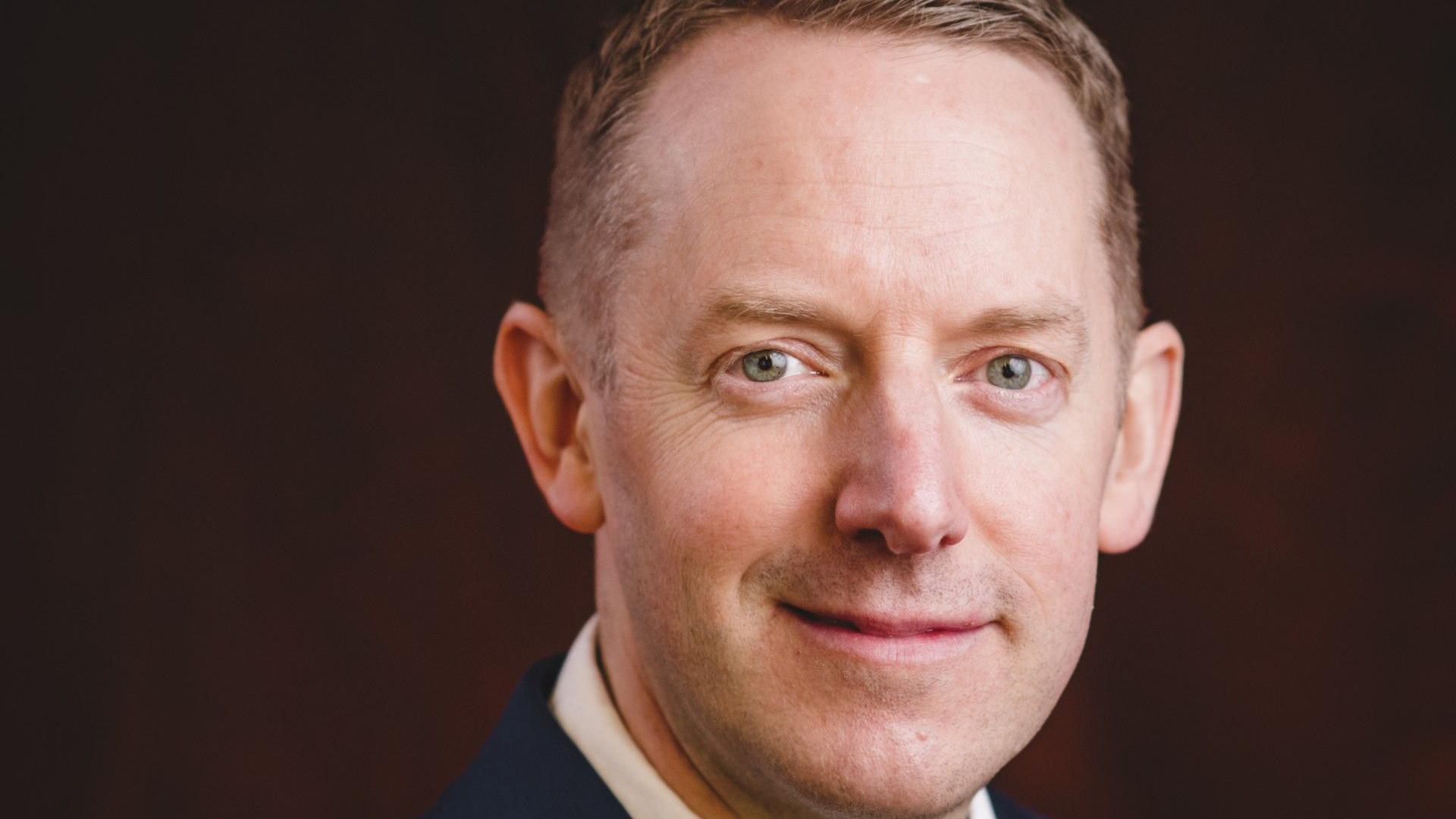 Close up of Mike Badley. Mr Badley is smiling at the camera. He is wearing a white shirt and navy suit jacket. He is sitting against a dark red backdrop.