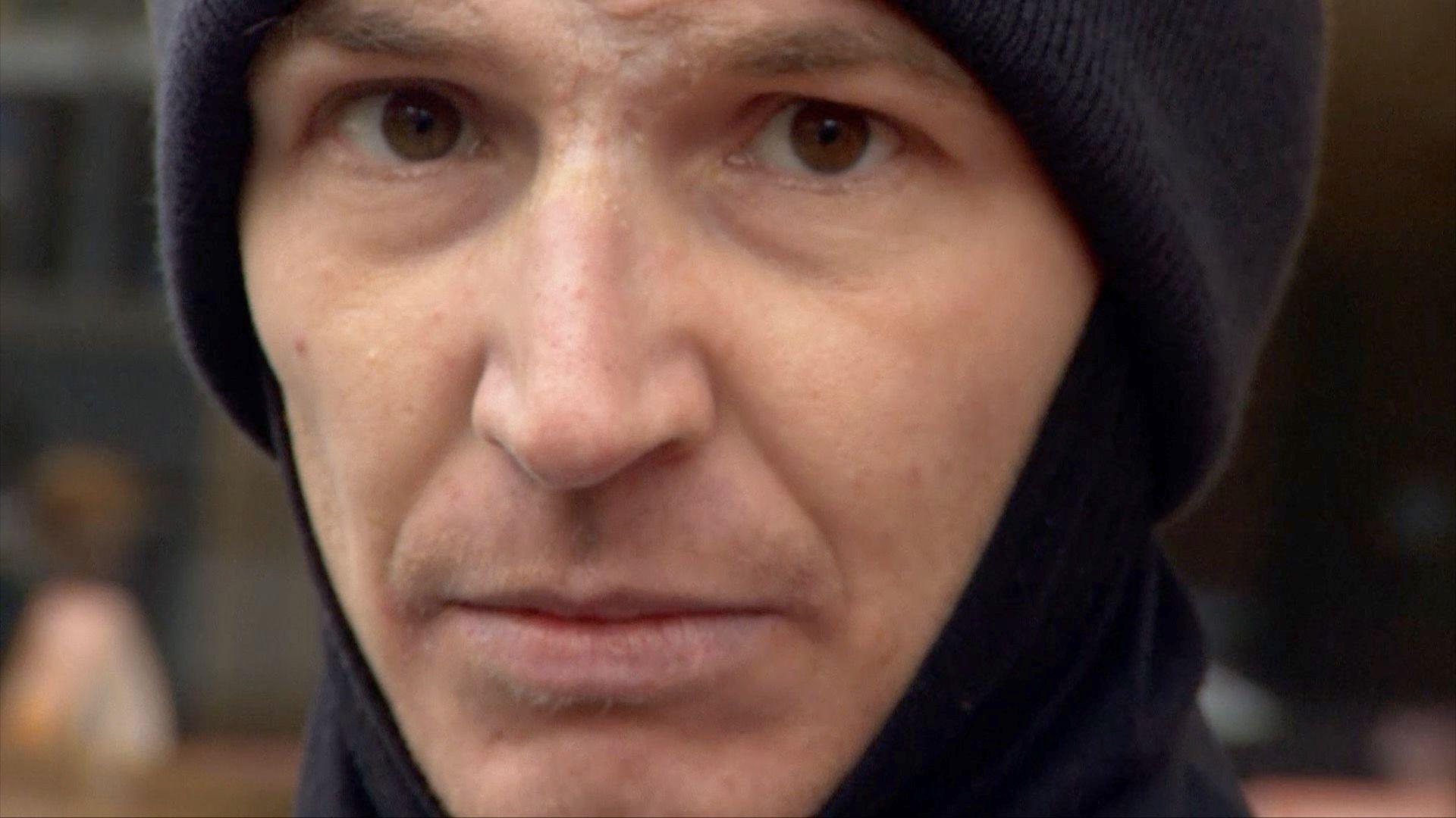 A close-up shot of man wearing a dark-coloured snood and a woolly hat. He is straight-faced and looking directly at the camera