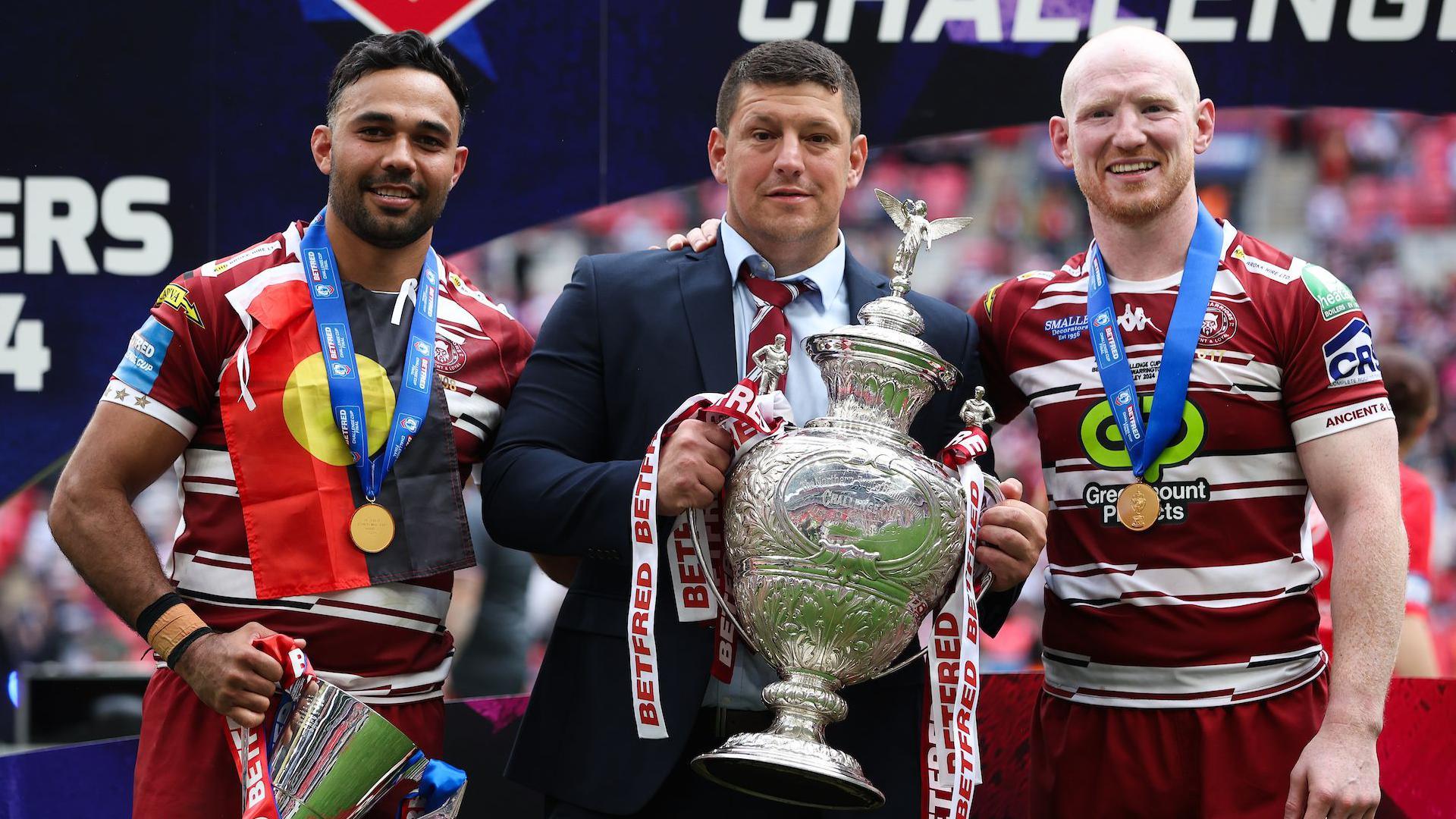 Bevan French, in a flag of the indigenous peoples of Australia and clutching the Lance Todd Trophy, stands with Matt Peet holding the Challenge Cup and Liam Farrell for a posed photo on the pitch at Wembley after Wigan beat Warrington in the Challenge Cup final.