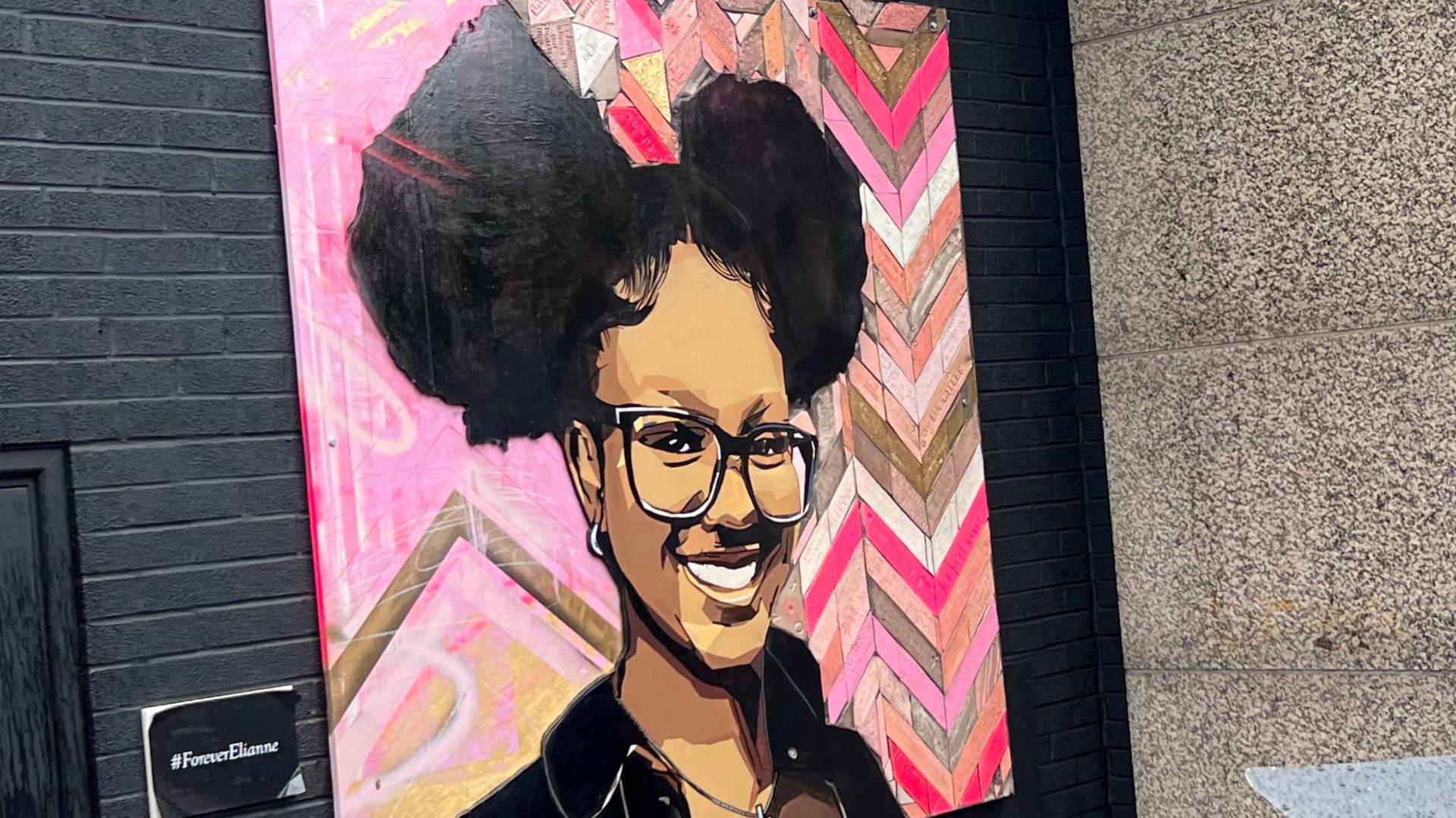 A mural of Elianne Andam has been painted on a wall in Croydon in south London. In it she is smiling and wearing glasses. Her hair is up. It shows her head and shoulders and the background is a pink design with a geometric pattern