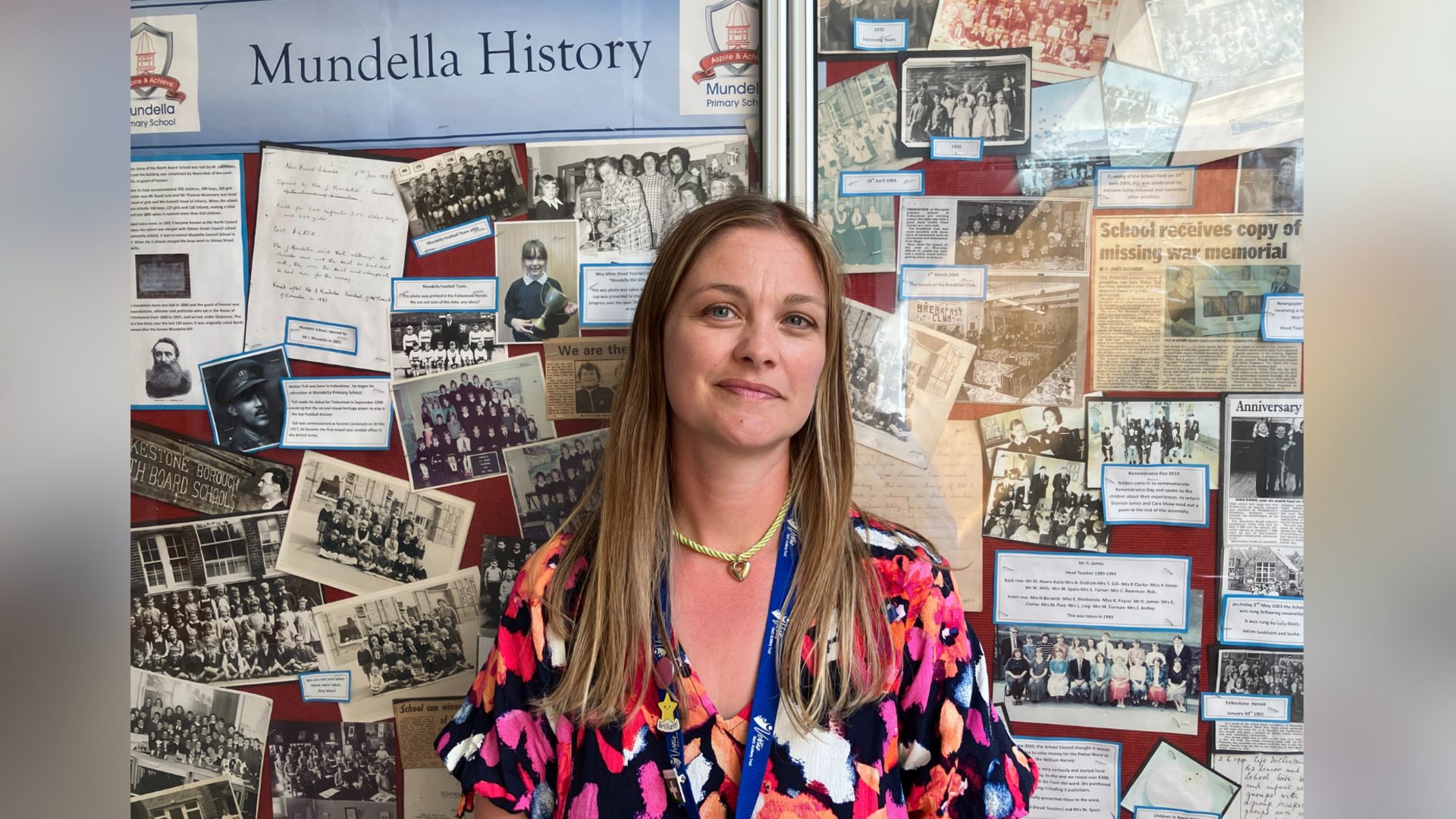 Co-headteacher of Mundella Primary School in Folkestone, Kent, Lauren Wharmby. Lauren is stood in front of a display wall dedicated to the school's history, and is wearing a patterned dress and blue lanyard. She has long blonde/brown hair.