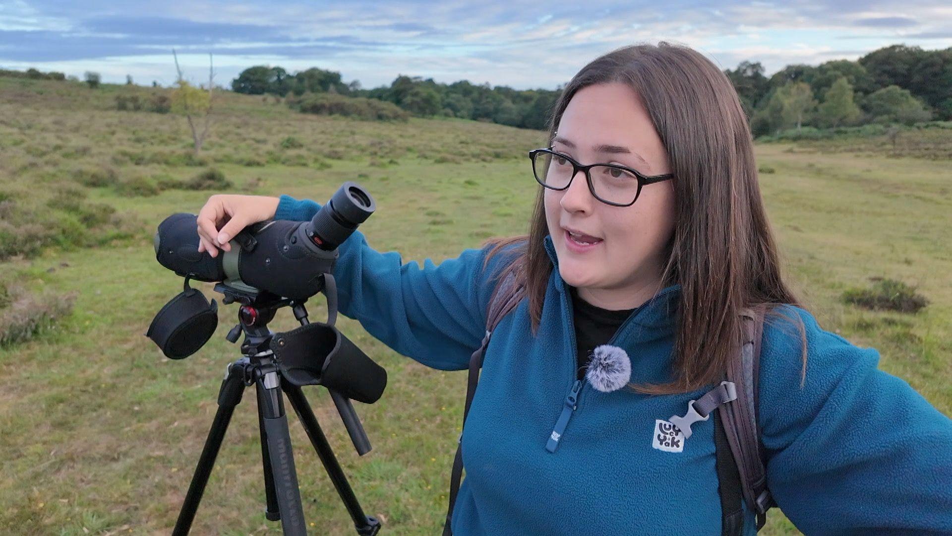 A young woman with dark hair and glasses stands next to a telescope on open heathland