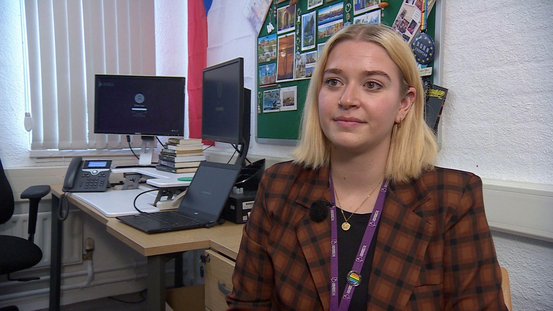 Veronika Carruthers, a woman with shoulder length blonde hair, a nose ring and wearing a checked brown blazer over a black top, and with a gold necklace and purple lanyard around her neck. She is sat in front of a desk with computer monitors