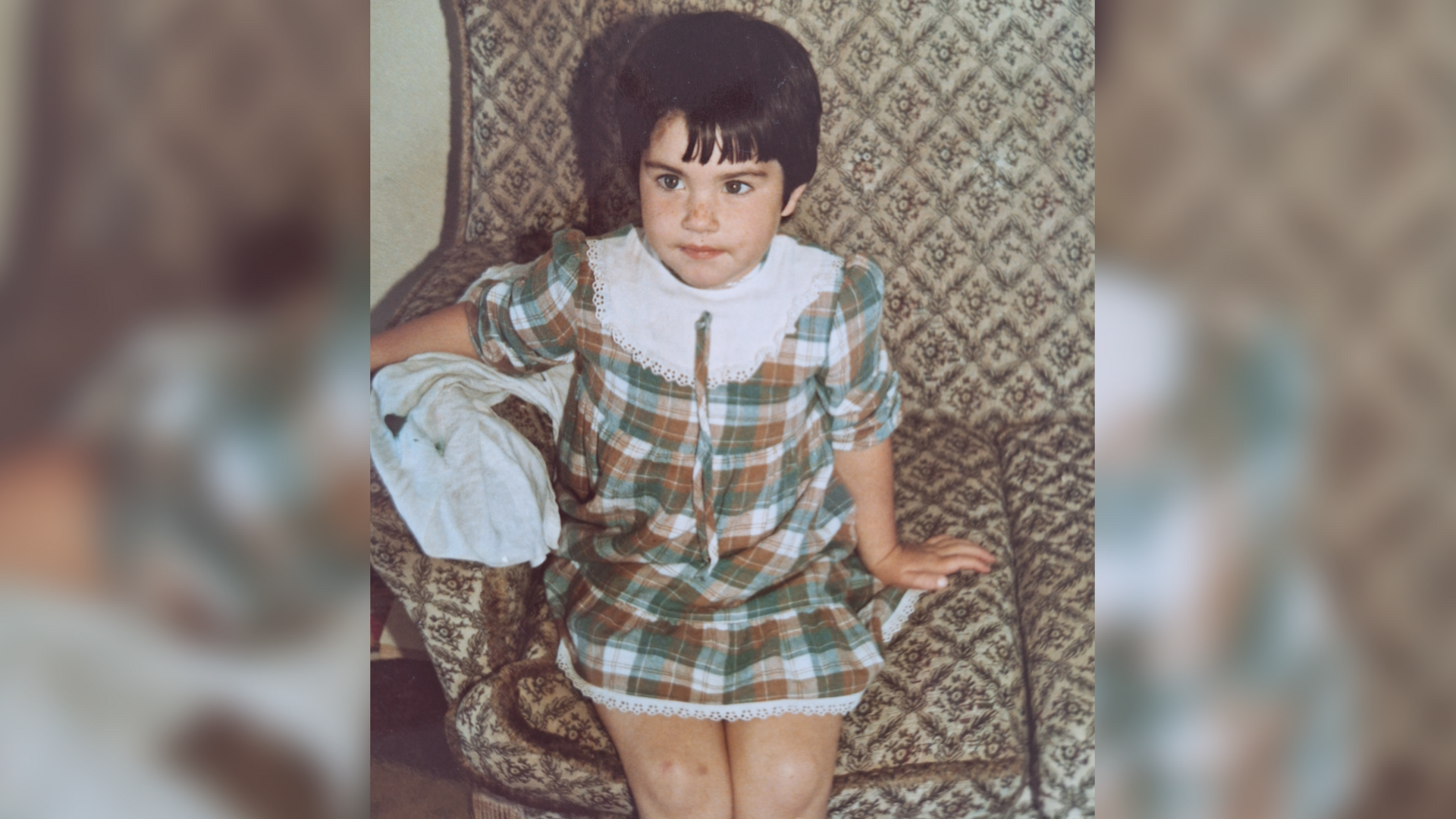 Rebecca aged about four years old sits on an old fashioned looking sofa, her arm over the arm rest. She is wearing a green and cream tartan dress which has a big white collar. The photo is from the 1980s. Rebecca has short dark hair, freckles on her nose and is looking past the camera at someone else in the room. 