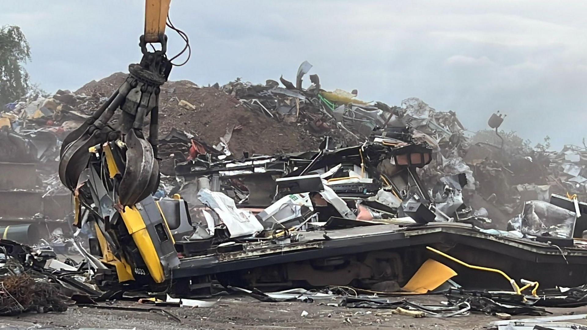 A scrap metal yard, with in the foreground the remains of what was formerly a Tyne and Wear Metro carriage, the front is held by a giant hydraulic grabber. In the background is a large pile of scrap metal.