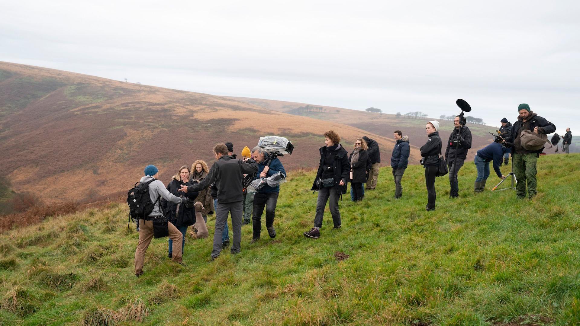 Stood on a hill sloping the left of the image, a crew of 18 people gathers around, some with boom mics and cameras. Behind them is a brown-coloured hill.