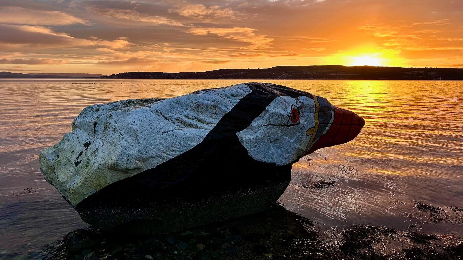 A dark rock with markings to make it look like a puffin. It has a colourful 'beak' with a dark eye painted slightly further up. It is in front of a body of water with a bright orange sun rising from the horizon in the background.