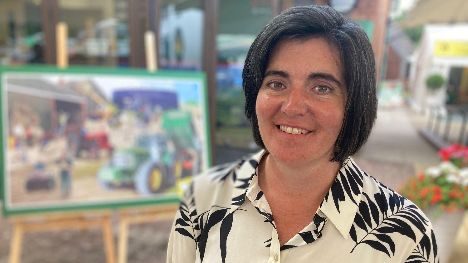 Abi Reader, president of NFU Cymru, smiles at the camera. She has dark, chin-length hair and wears a white blouse with black leaves on it. 