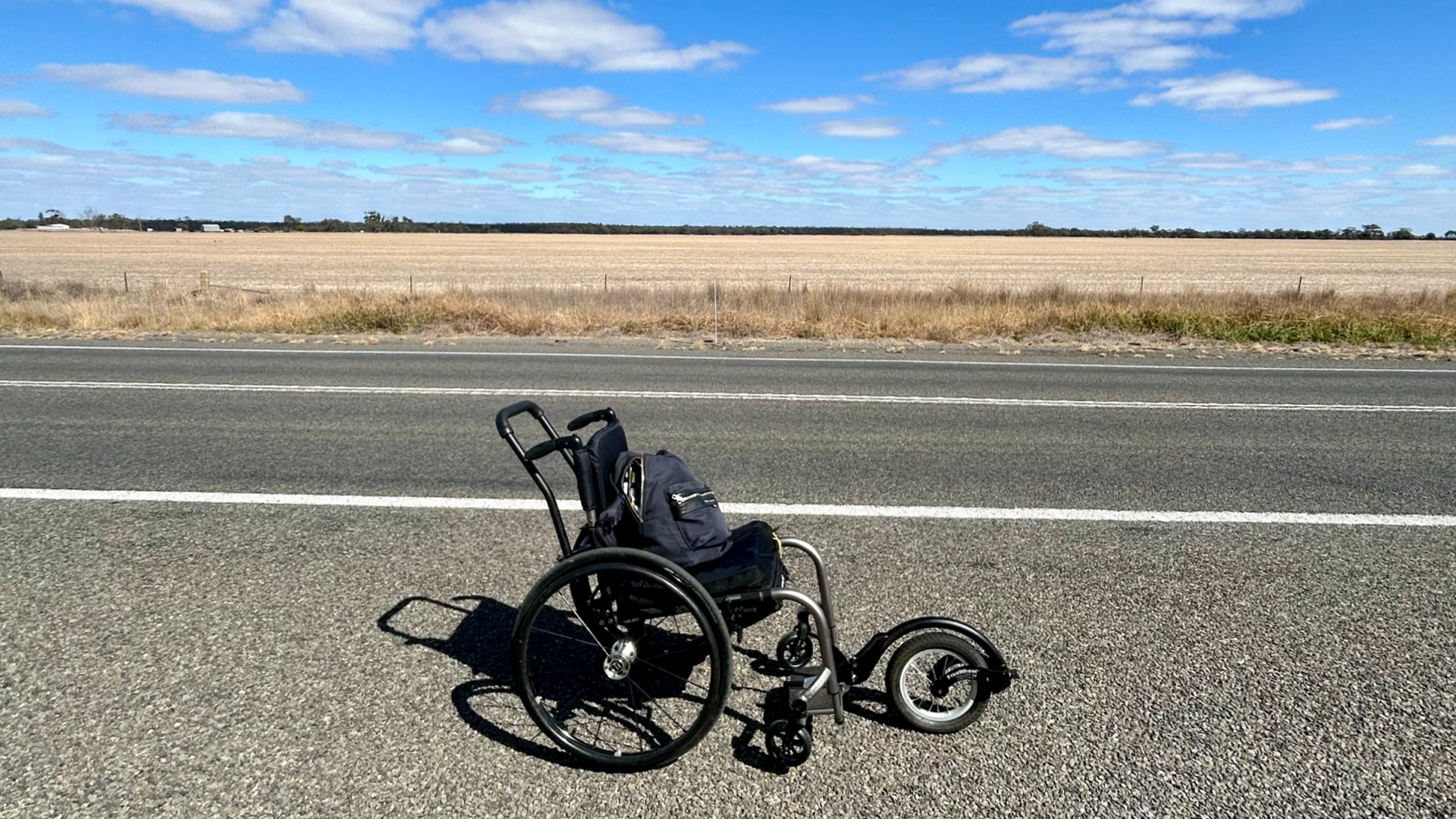 A wheelchair is parked on an empty straight road. In the background there is vast Australian landscape and blue skies.