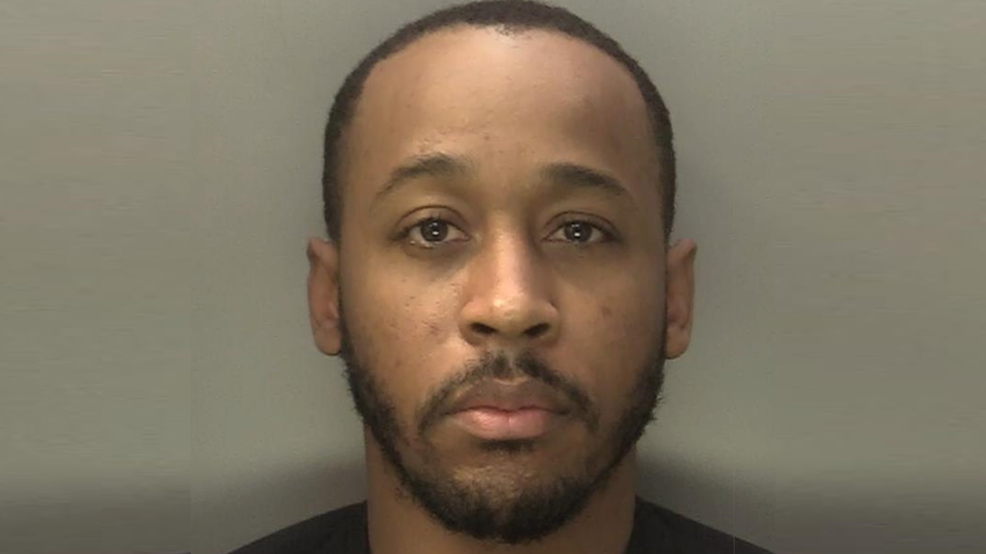 A police mugshot of a man with short dark hair and a short dark beard and moustache, wearing a black T-shirt, stood against a plain wall.
