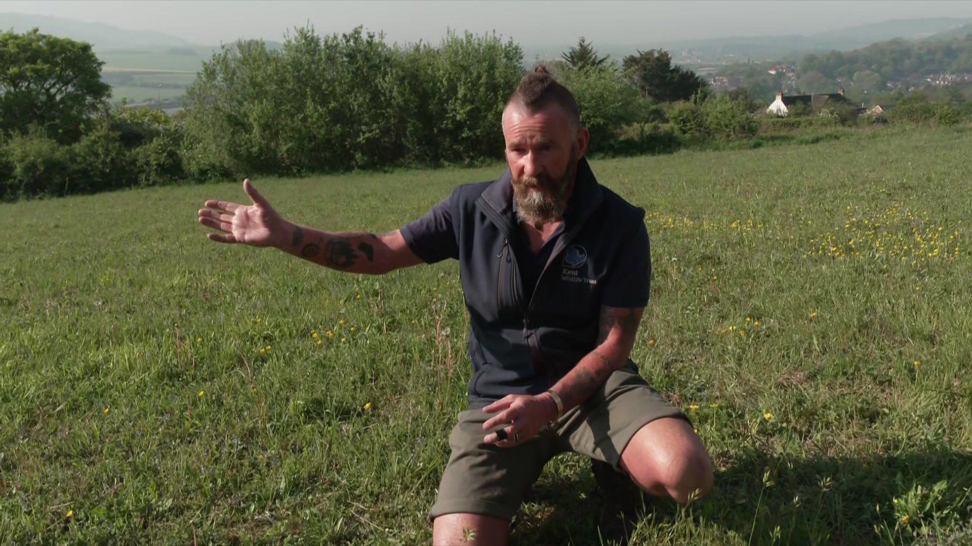 A man in shorts and a gilet points at a field, which he is kneeling in. It is very sunny.
