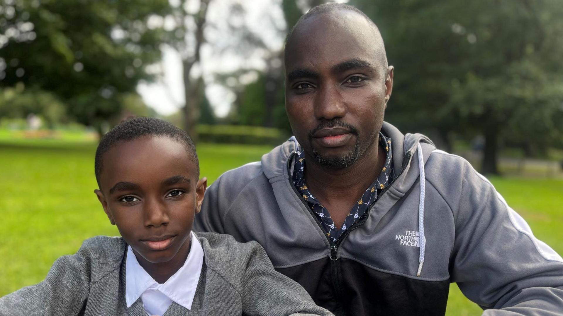 A man and child in a park. The boy is wearing a school uniform and the dad is wearing a shirt and a sports hoody