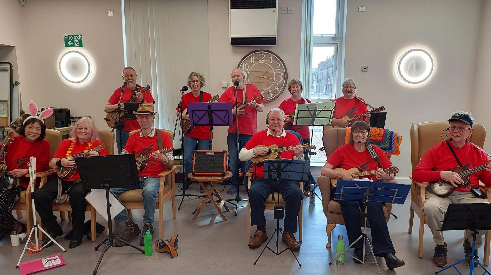 A room full of ukulele players all holding their instruments. The front row is made up of six players all sat in armchairs with music stands in front of them. The second row are stood behind the with microphone stands beside them.