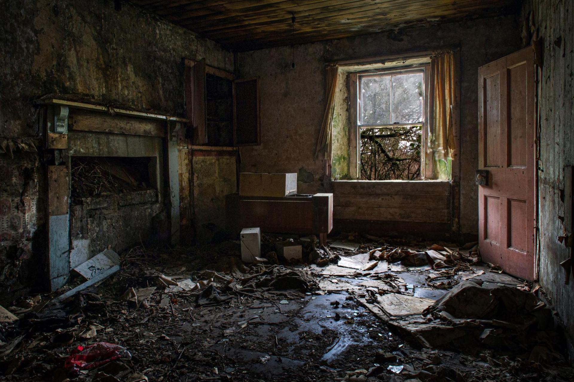 Plaster and rubbish covers the floor of the room. There is a window with most of the glass missing and red wooden door that is ajar. An old fireplace is partially blocked up.