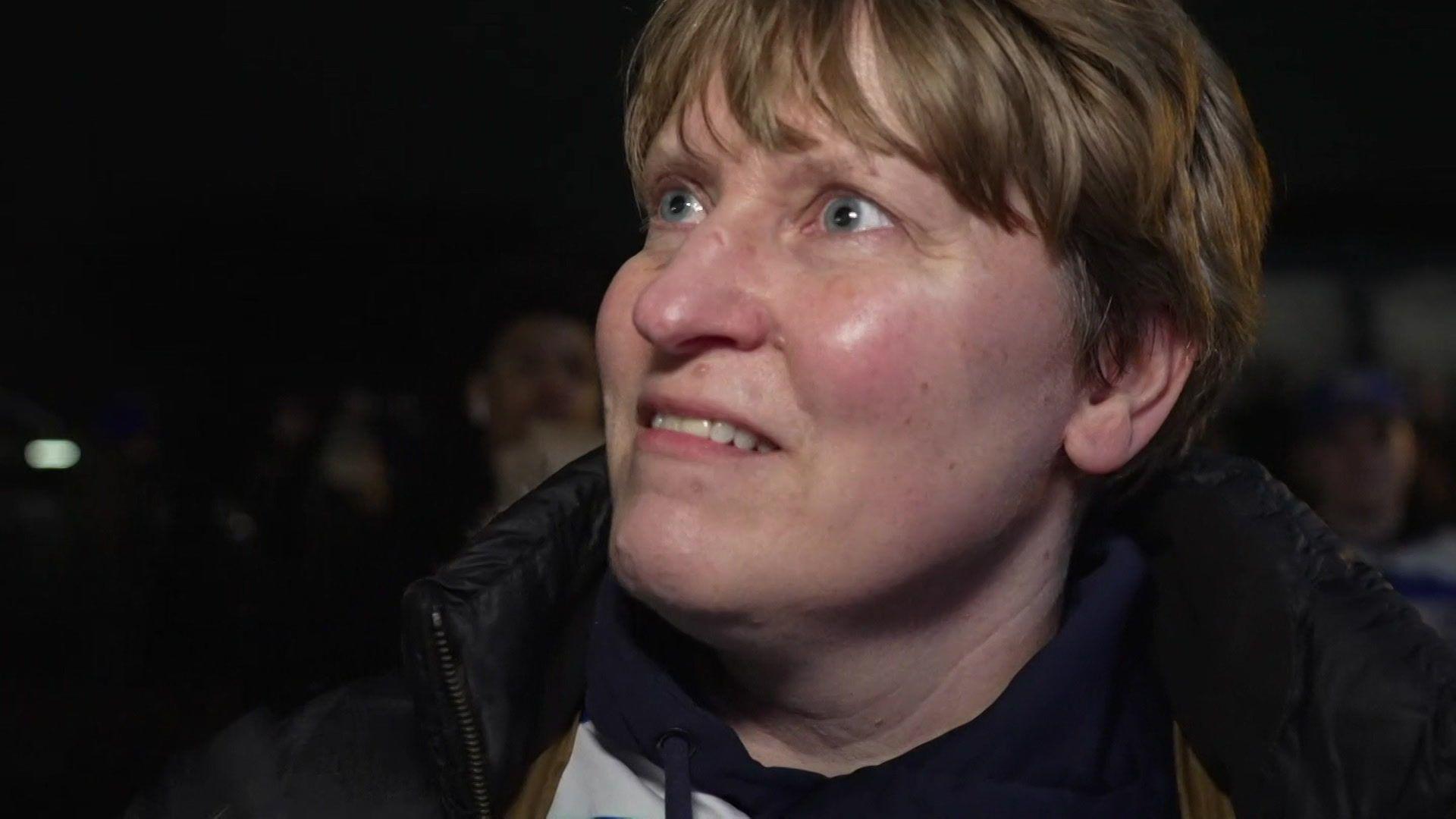 A head and shoulders photo of a woman with short blonde hair, wearing a black coat. She's looking slightly past the camera. It's dark and there's a crowd of people behind her