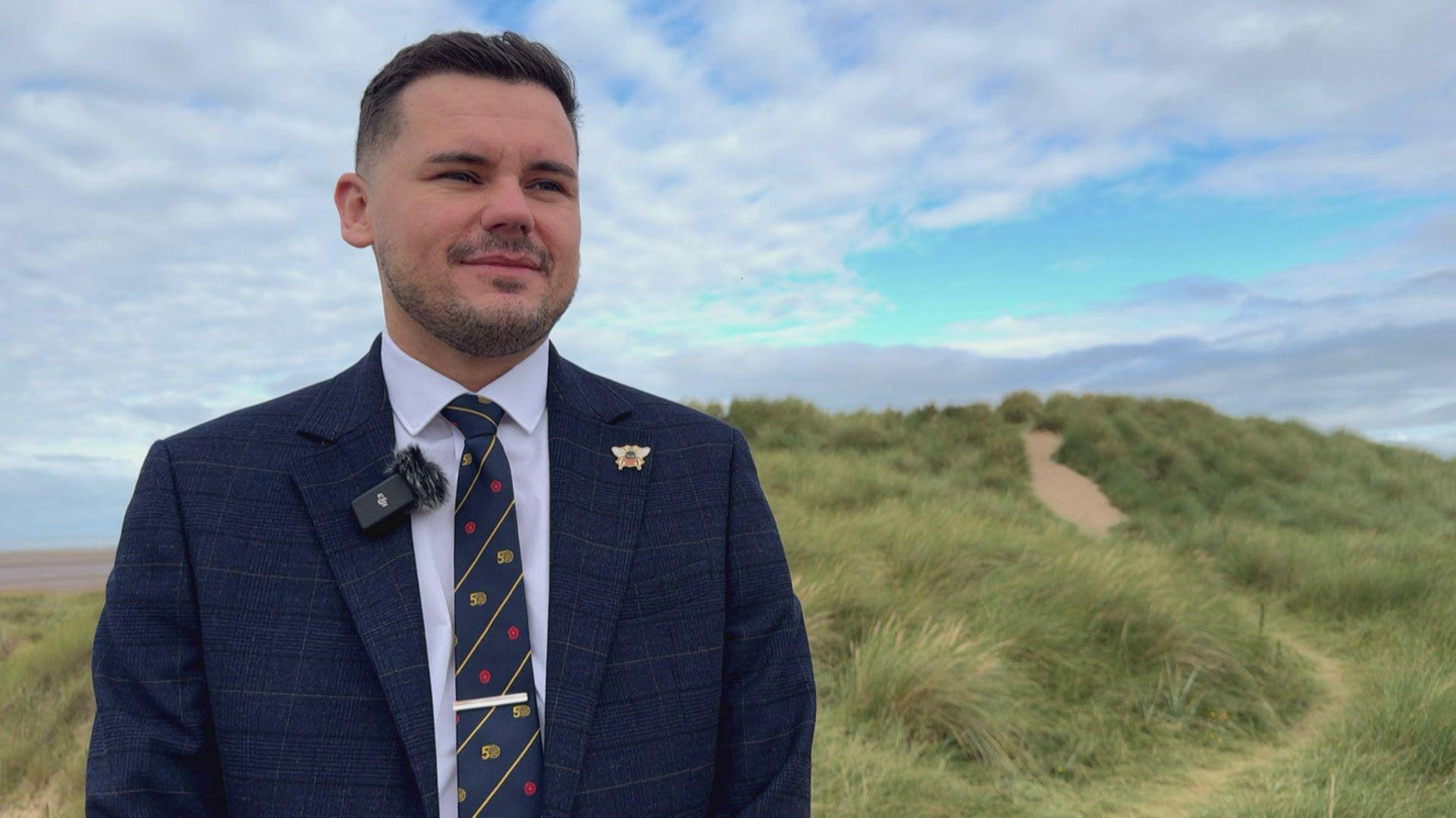 Lancashire County Councillor Joshua Roberts on the sand dunes at Lytham St Annes. The Reform UK politician is wearing a dark blue suit and tie. He has dark hair and is smiling.