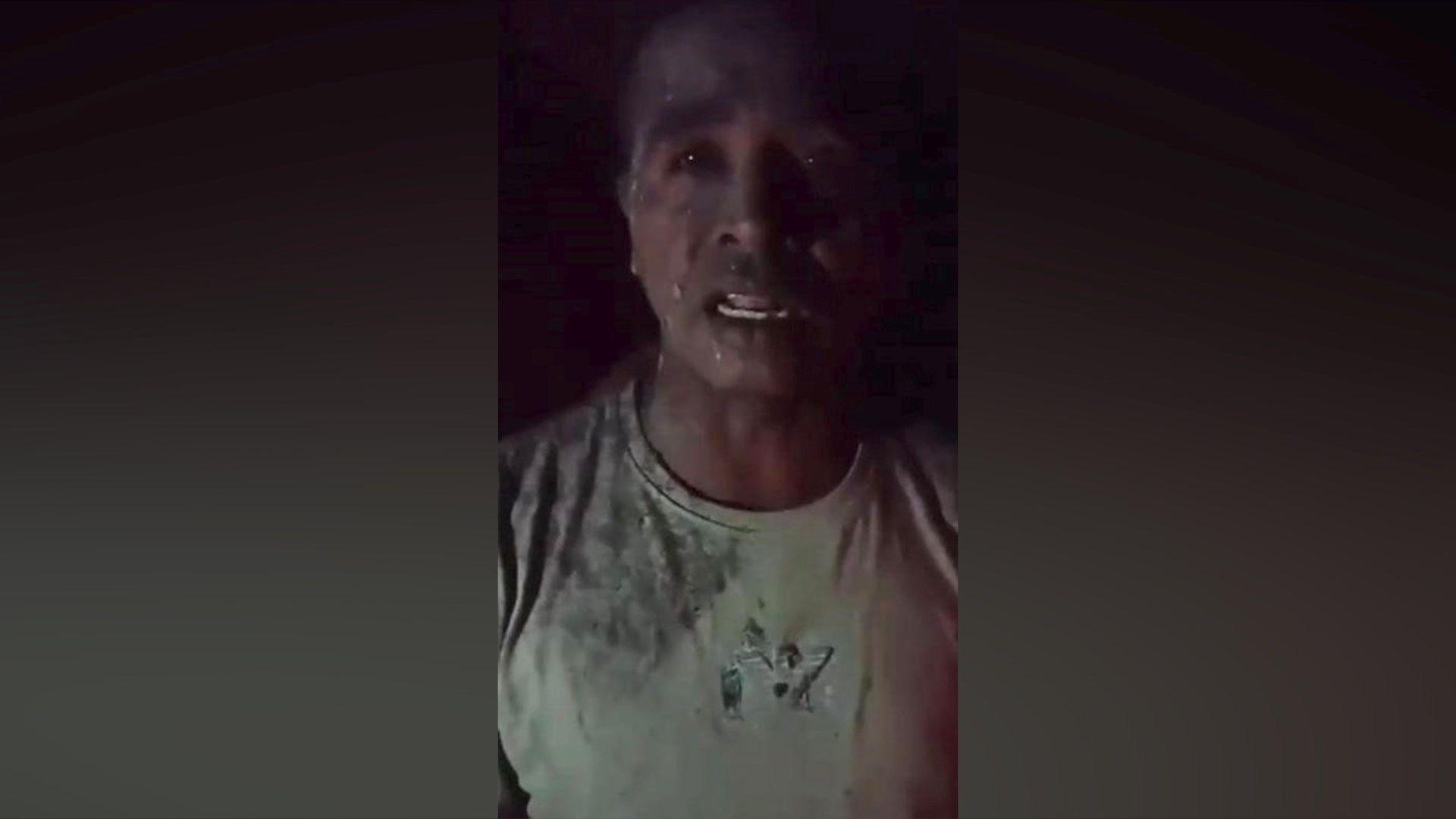 Ash covers a distressed-looking man's T-shirt, while bits of volcano ash are spattered on his face in a screenshot from a video.